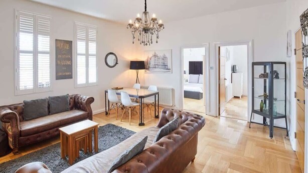 A spacious living room with a brown leather sofa, a matching armchair, a dining table with chairs, and a chandelier. - Host & Stay | Boutique Balcony (Whitby)