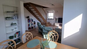 Ein Essbereich mit Holztisch, Stühlen und einer Vase mit grünen Früchten. Eine Treppe führt in einen weiteren Raum mit einer Couch und einem Bücherregal.