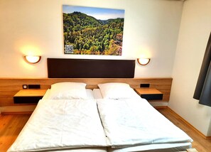 Habitación de hotel con una cama, dos mesitas de noche, un cabecero y un cuadro colgado en la pared que muestra un paisaje de bosque.