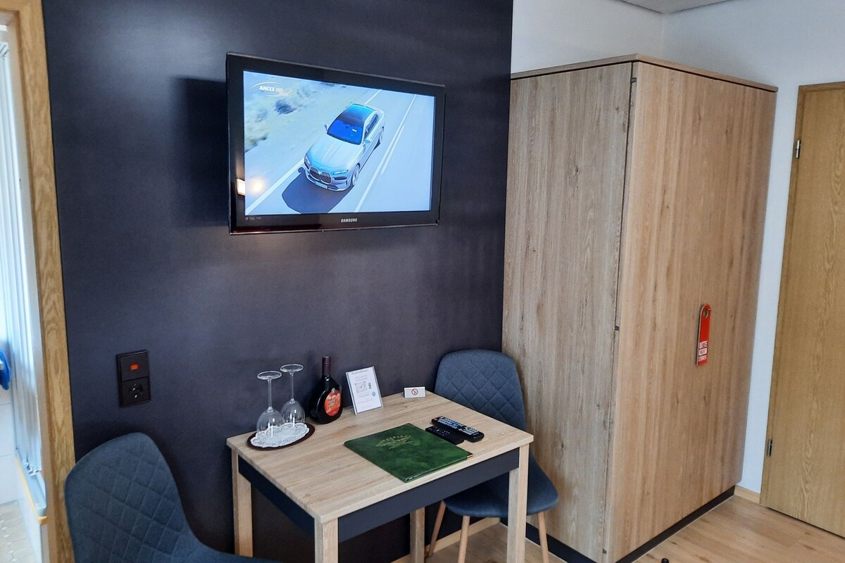 Una habitación con un televisor en la pared mostrando un coche, una mesa con un mando a distancia, gafas y un certificado. Hay dos sillas y un armario.