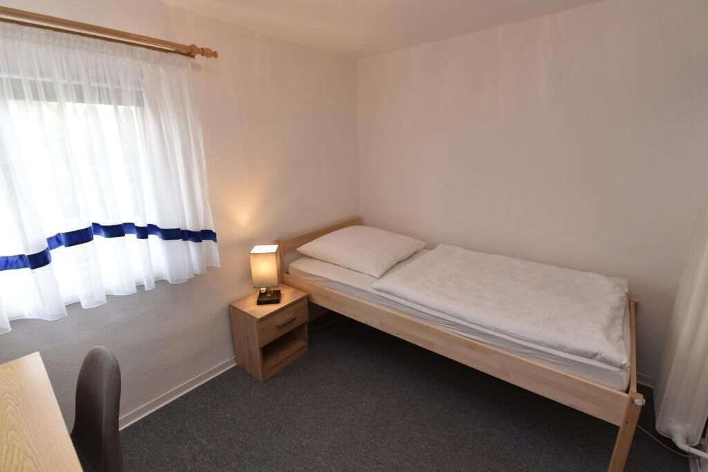 A small, single-bed room with a wooden frame, a bedside table with a lamp, and a chair.