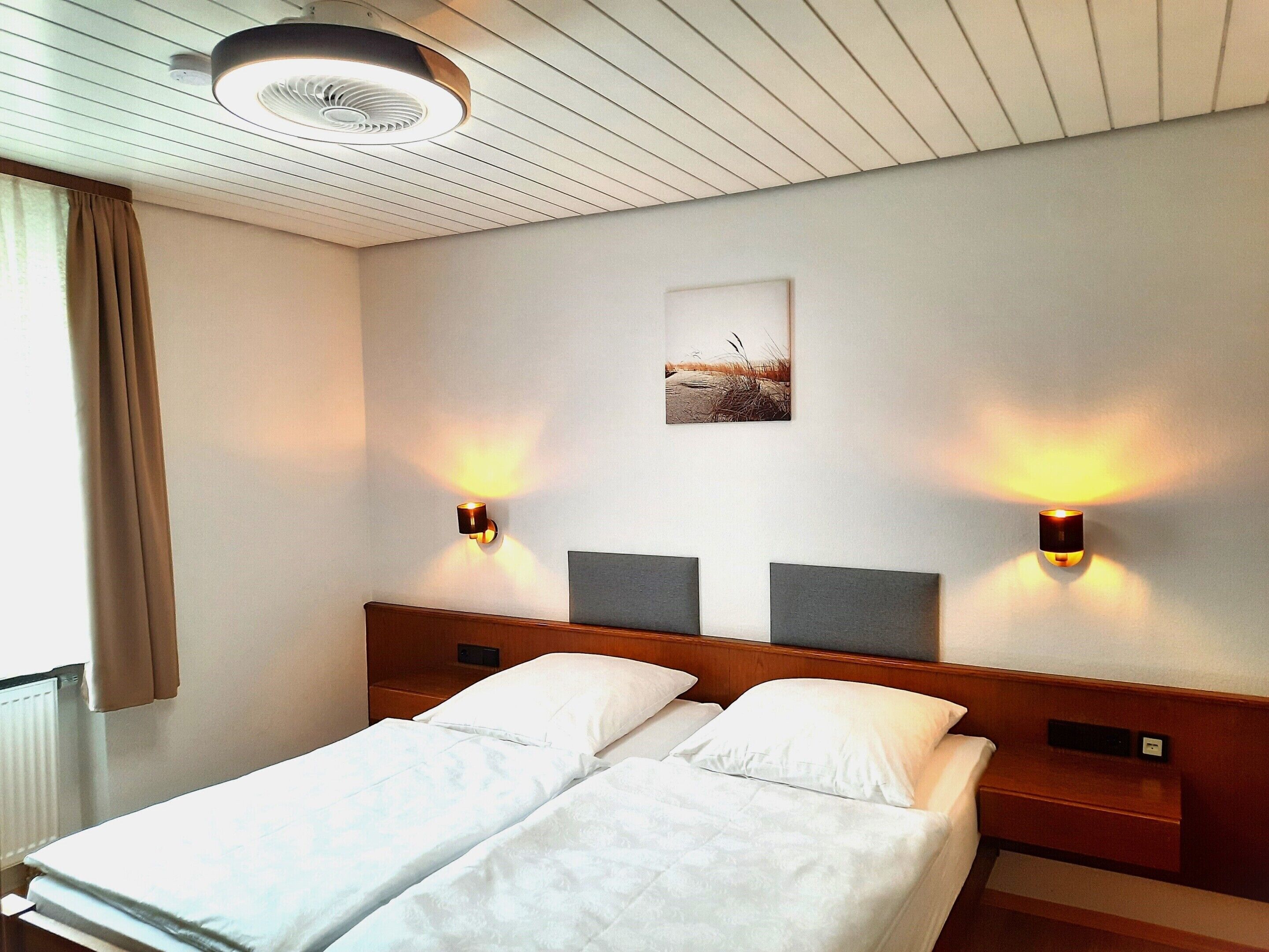 A hotel room with two beds, a ceiling fan, wall-mounted lights, and a picture on the wall.
