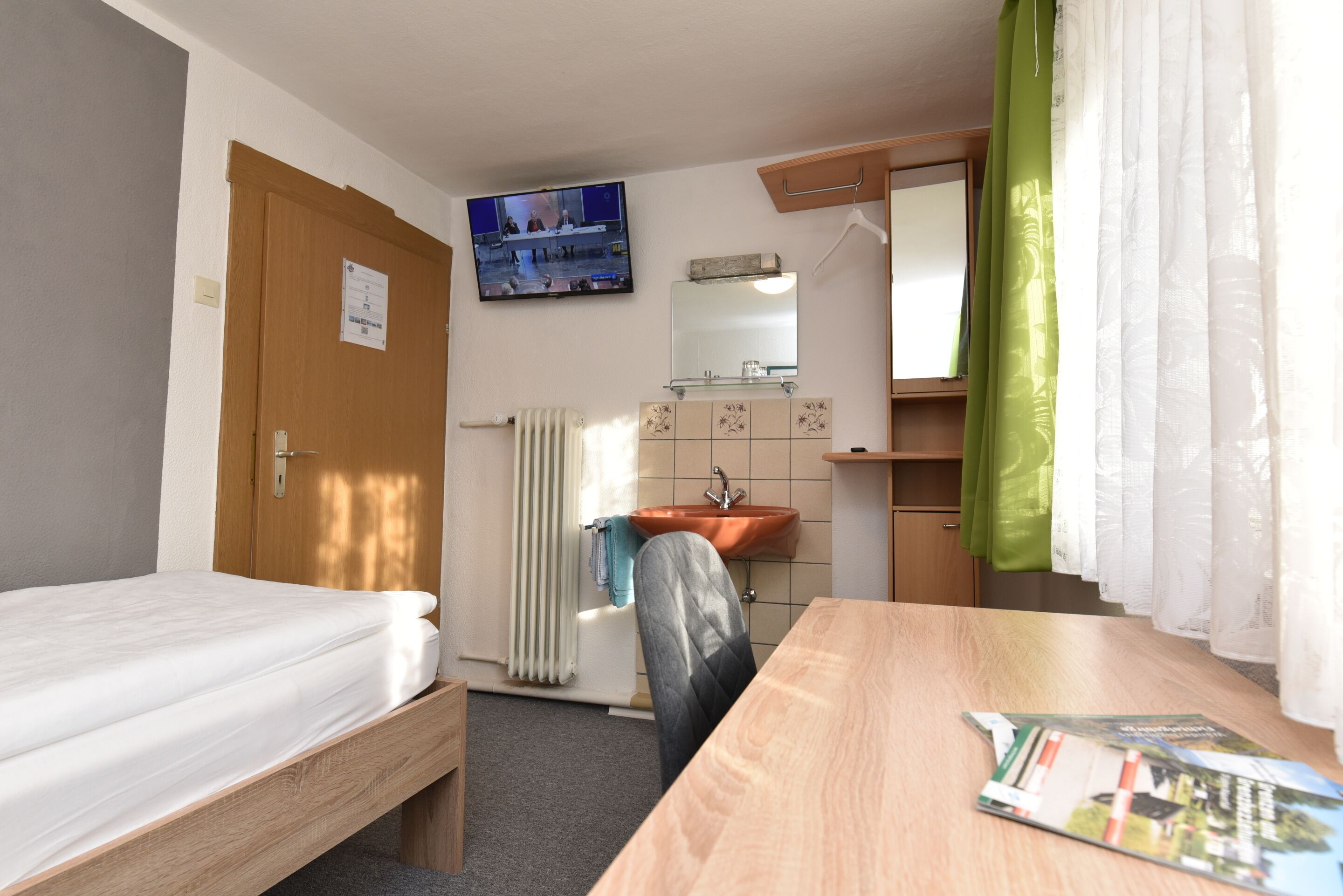 Habitación de hotel con una cama individual, un escritorio de madera, un espejo, un lavabo y un televisor montado en la pared.