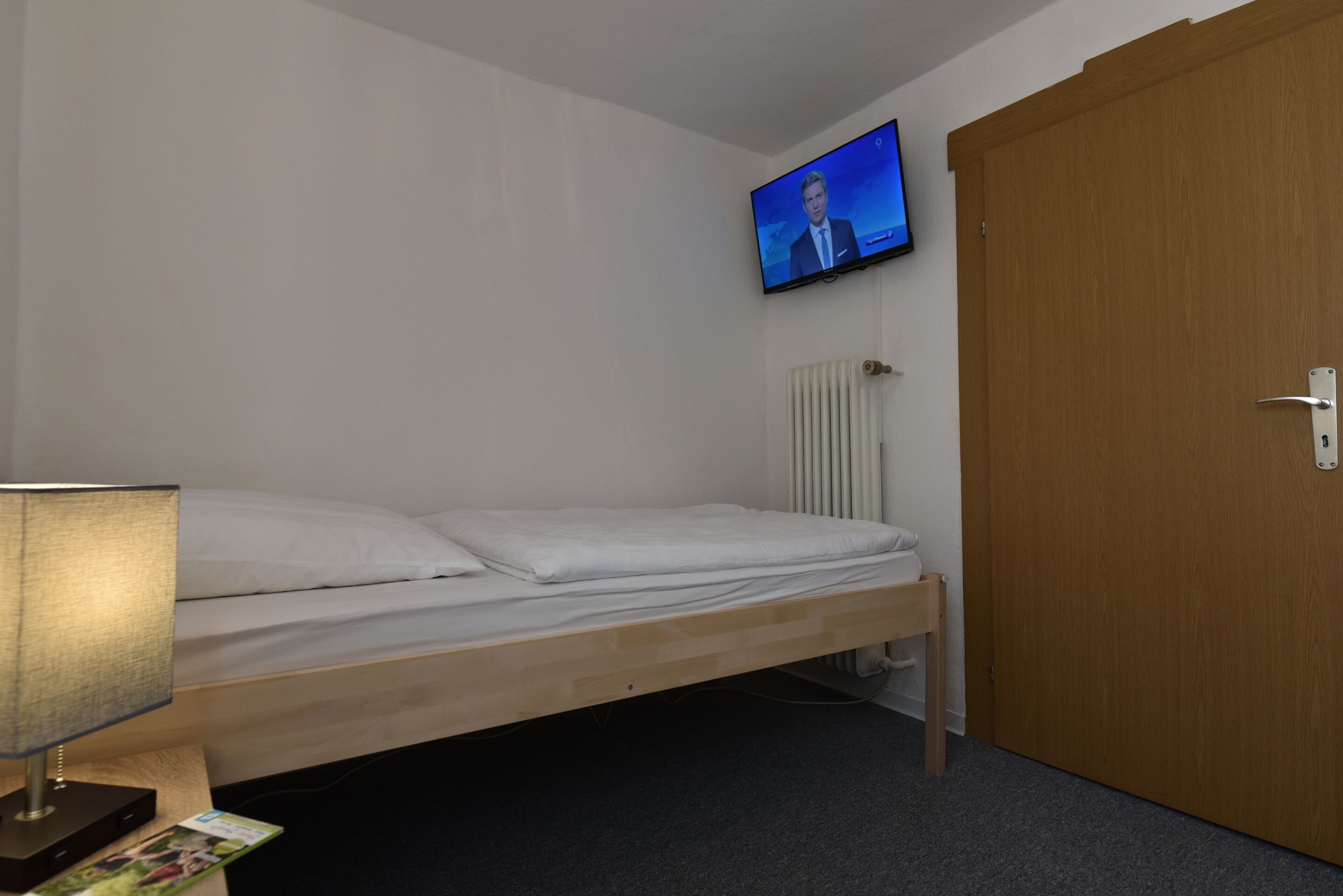 A single bed with a wooden frame, a bedside table with a lamp, a television mounted on the wall, a radiator, and a wooden door.