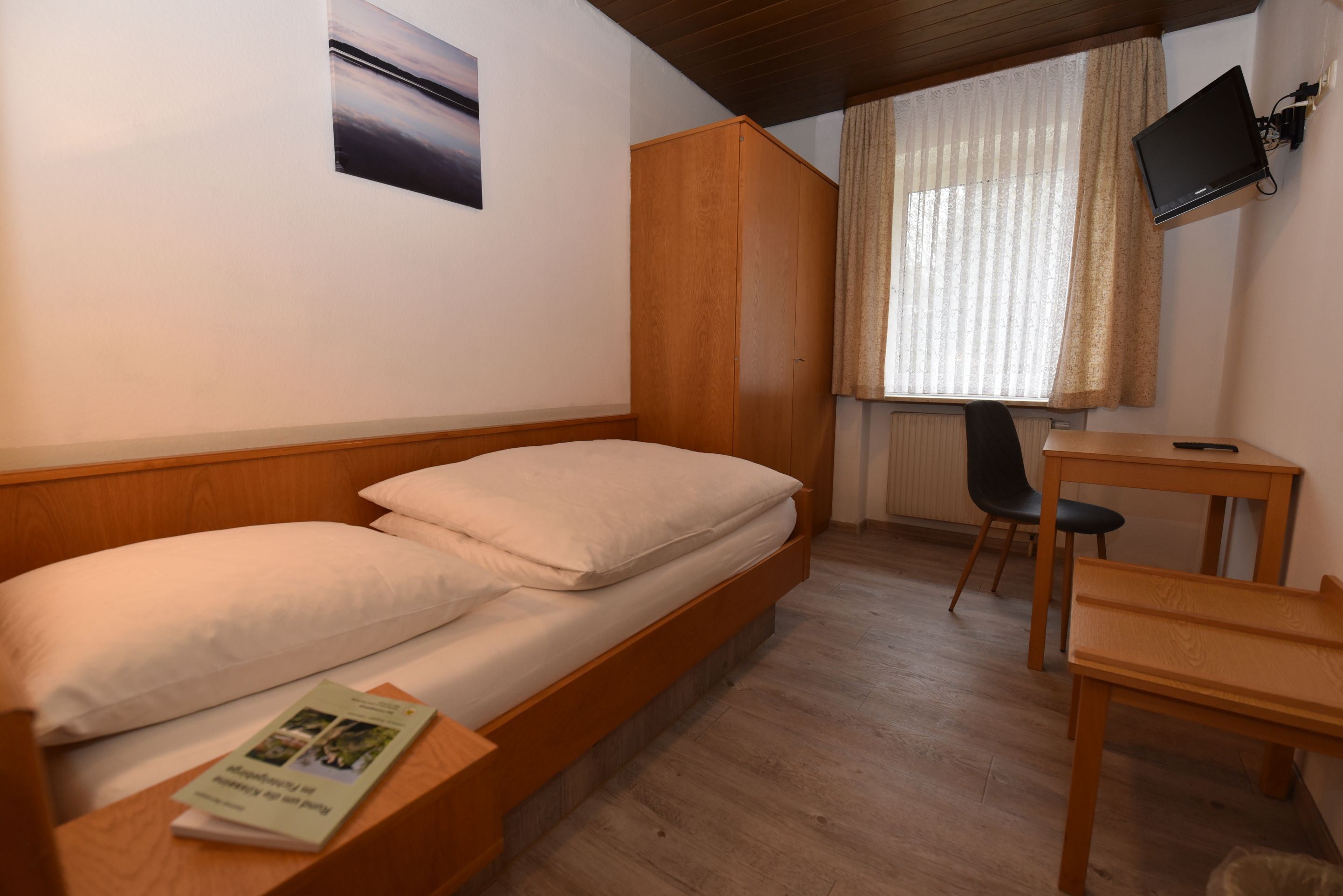 A single bed with a wooden headboard, a small desk with a chair, a television mounted on the wall, and a window with curtains.