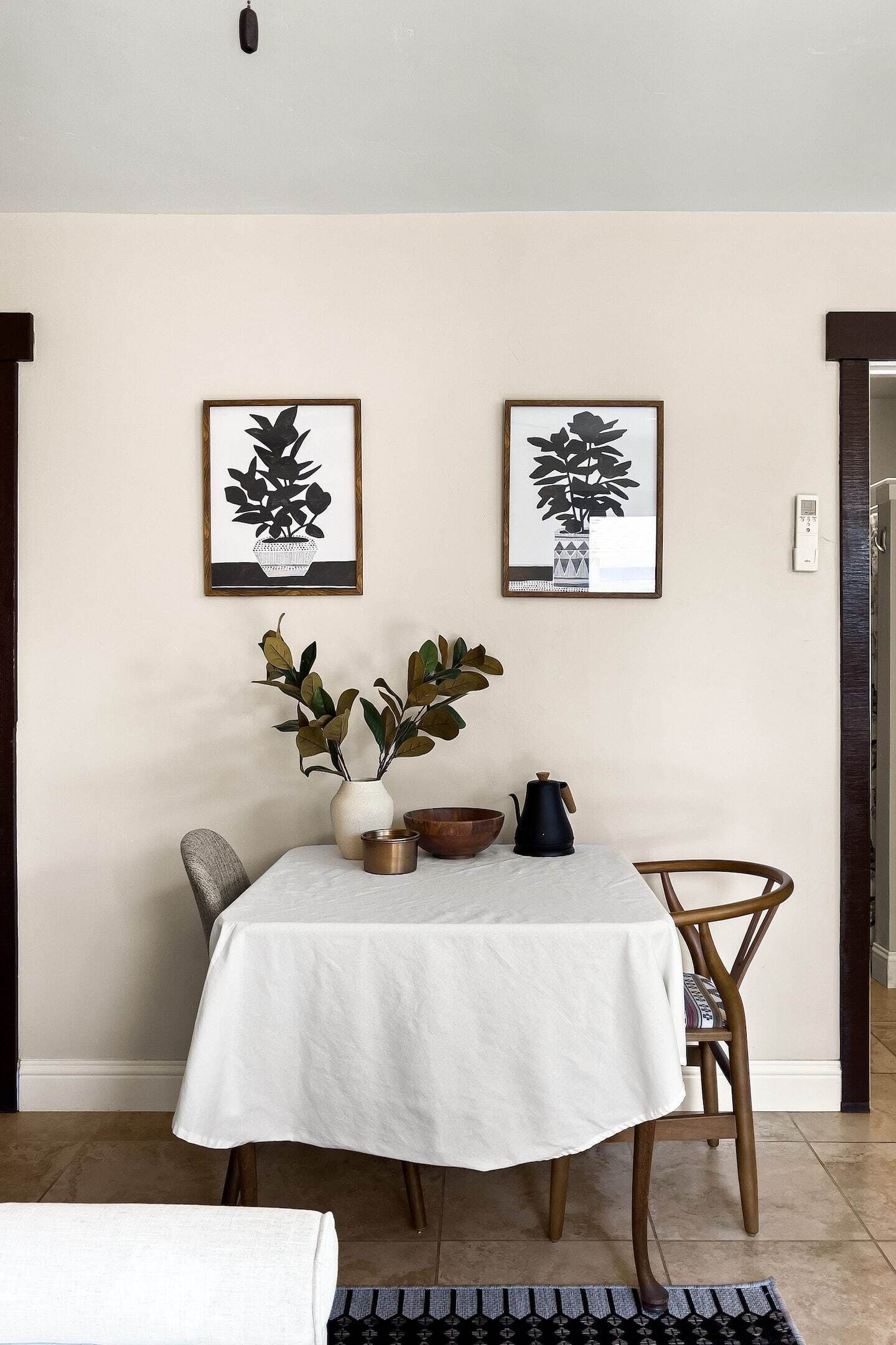 A dining area with a table set for two, a vase with green leaves, and two framed black and white plant illustrations on the wall.