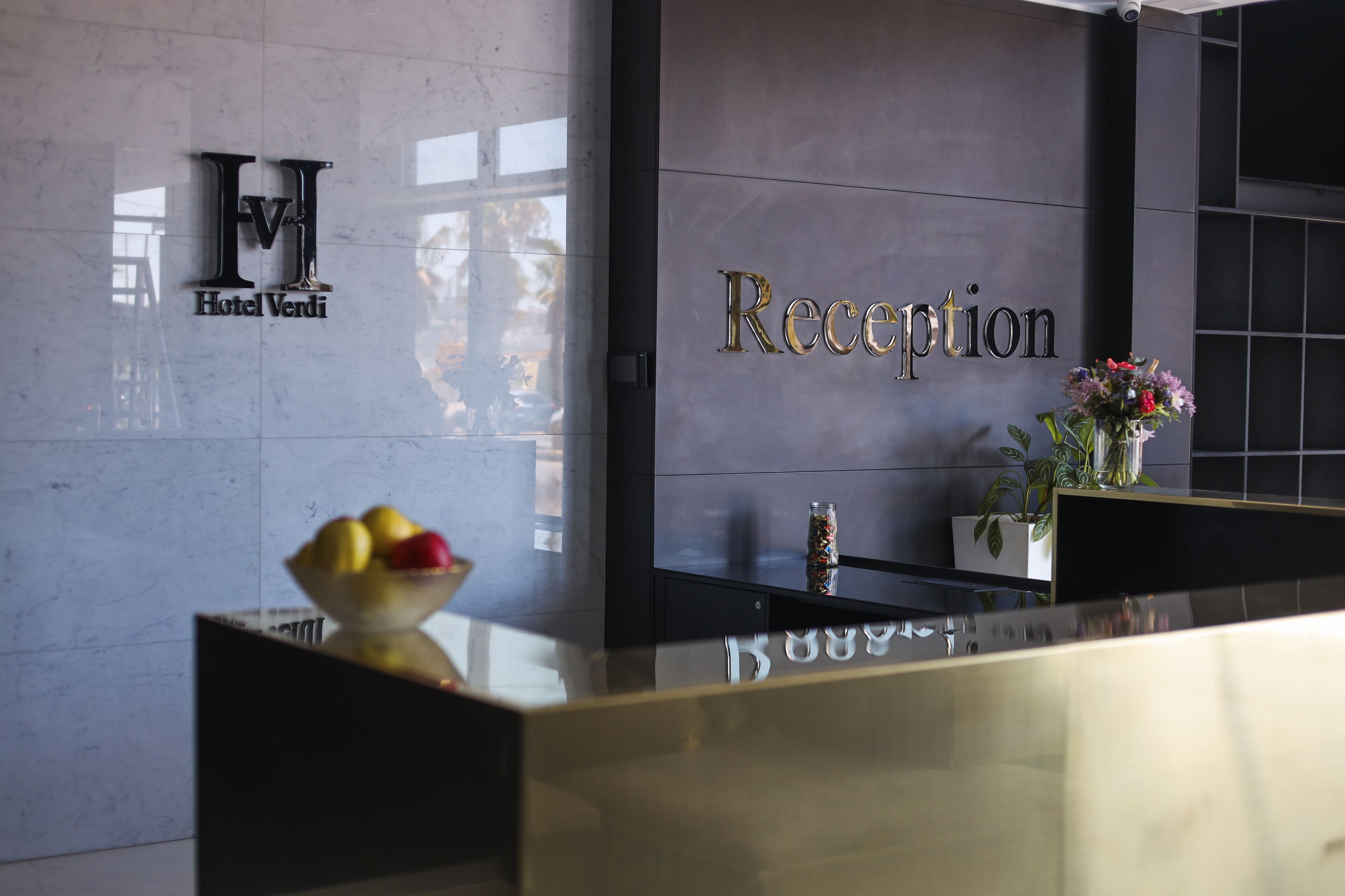 Een receptiebalie met een marmeren aanrecht, een schaal fruit en een vaas met bloemen. De achtergrondmuur heeft een bord met 'Hotel Verdi' en een bord met 'Receptie'.