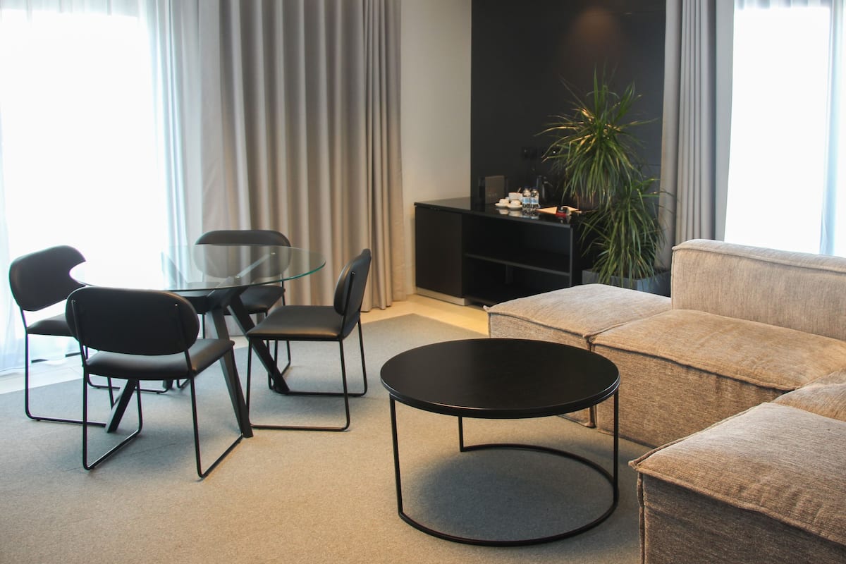 A modern living room with a round black coffee table, a beige sofa, and a glass dining table with black chairs. There is a black entertainment unit against the wall and a large potted plant.