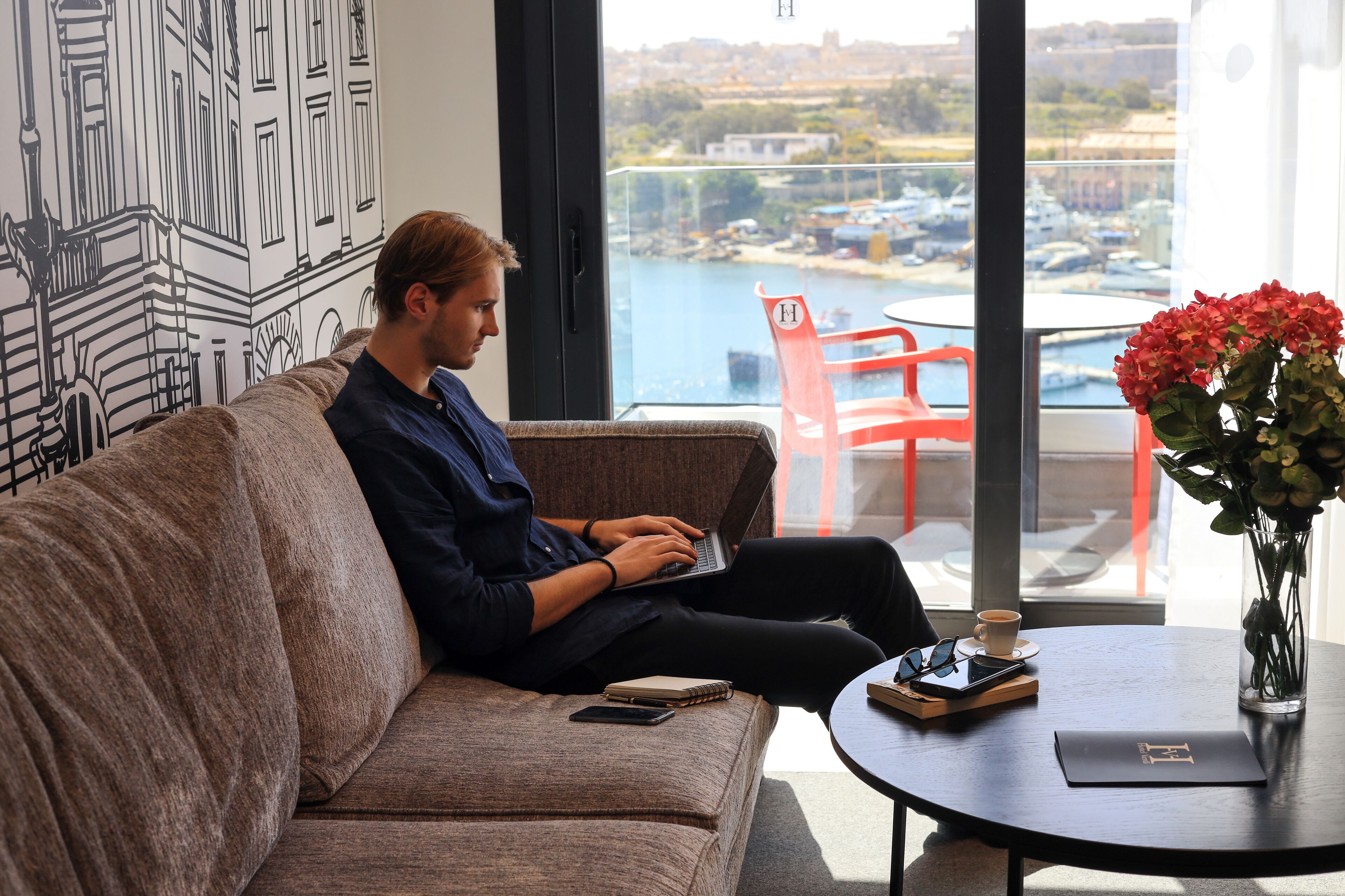 Eine Person sitzt auf einer Couch und benutzt einen Laptop, mit Blick auf einen Yachthafen durch große Fenster.