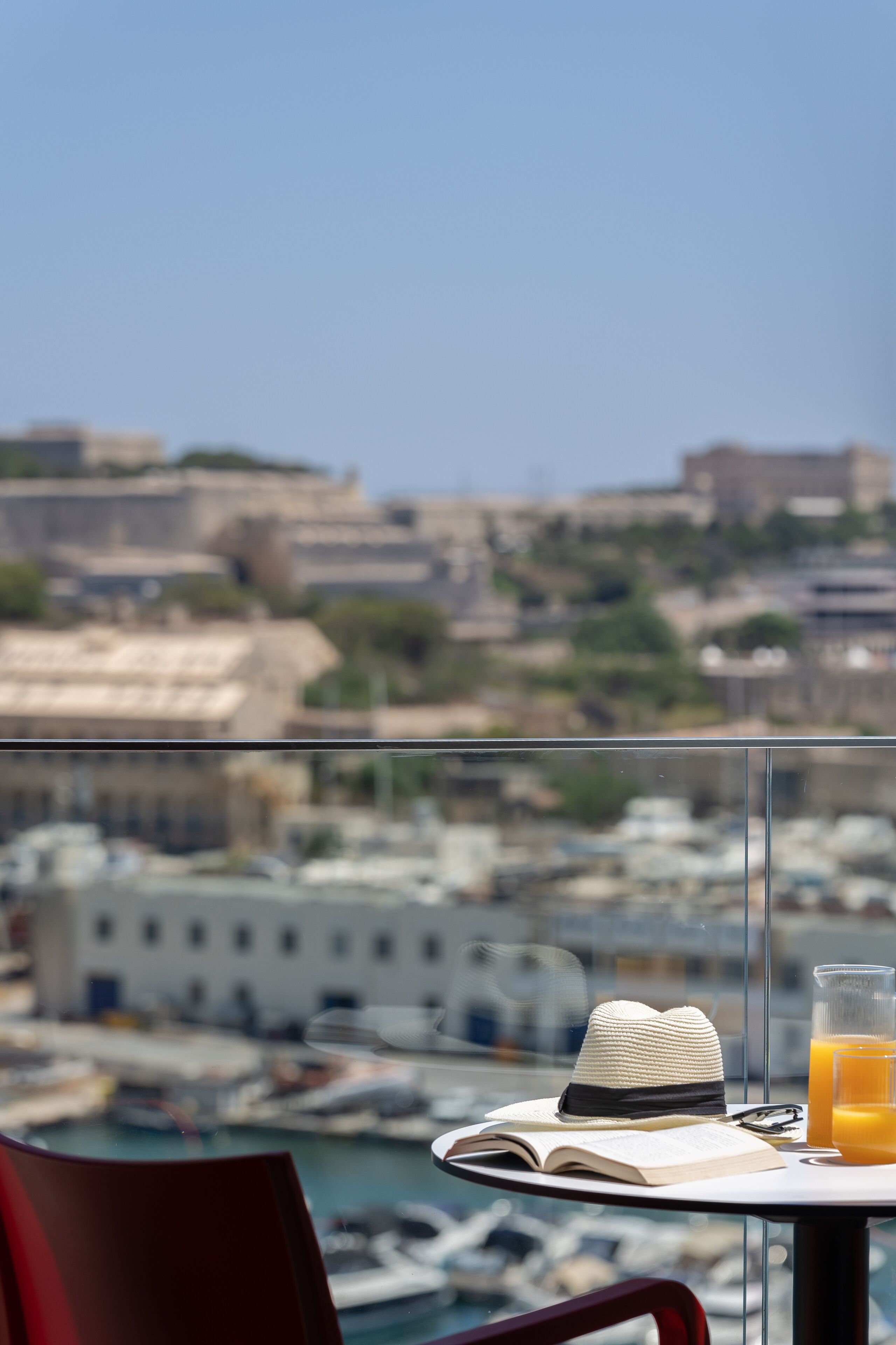 A table with a hat, book, and glass of orange juice, overlooking a marina with boats and buildings.
