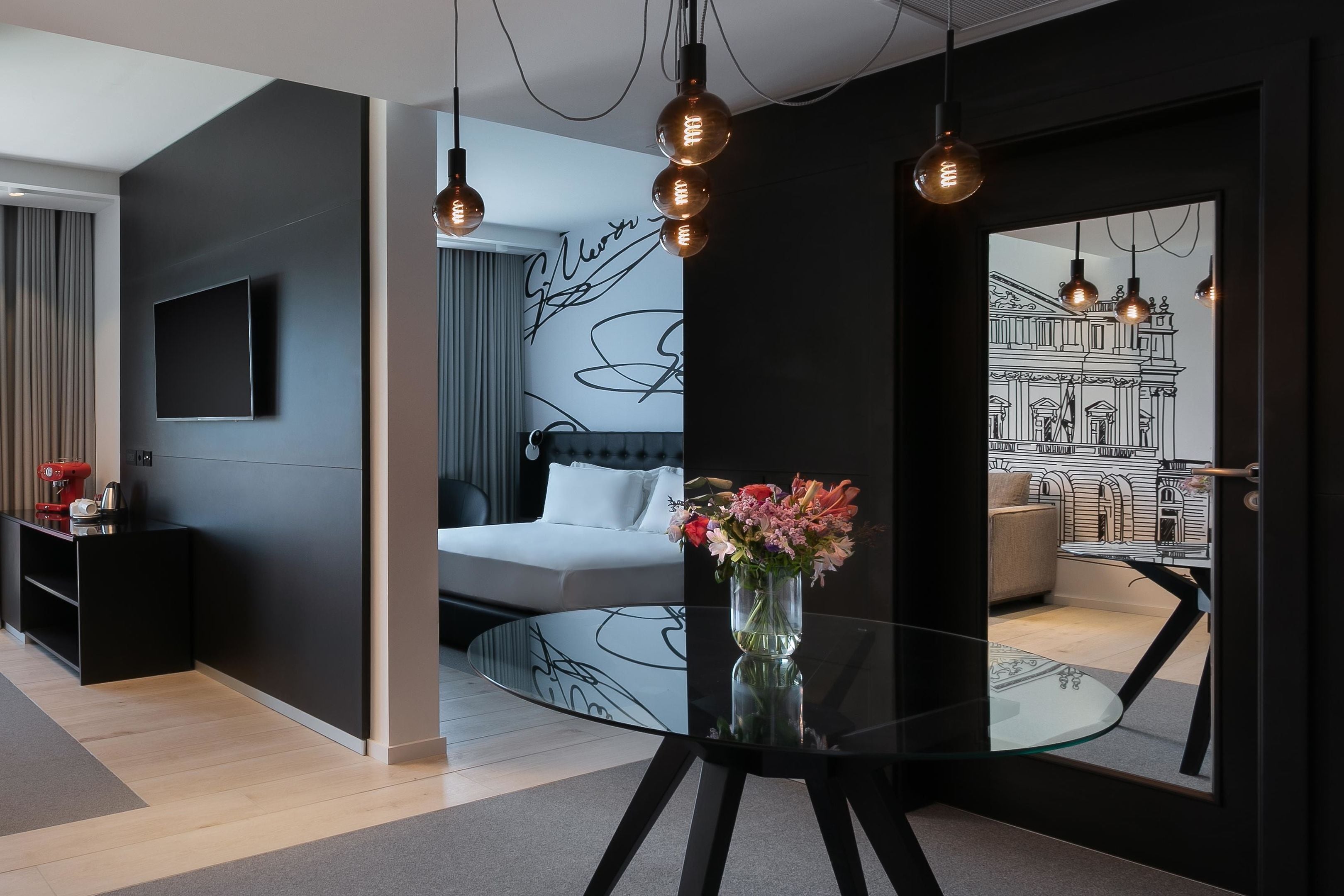 Een moderne hotelkamer met een bed, een glazen tafel met een bloevas, een flatscreen televisie en een muur met een zwart-wit muurschildering.
