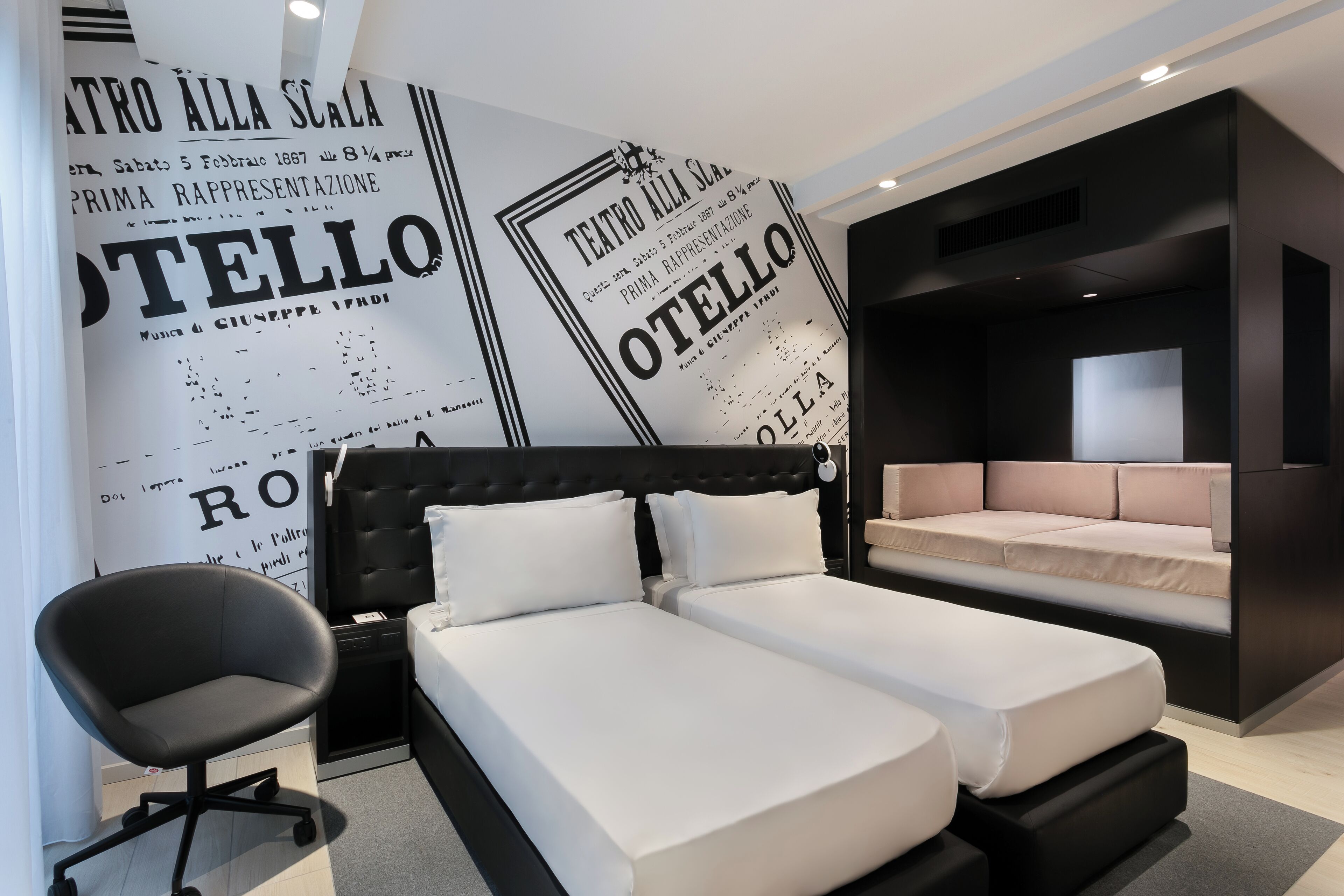 Camera d'albergo moderna con testiera imbottita nera, due letti con lenzuola bianche, una sedia da ufficio nera e una parete ampia con testo in bianco e nero.