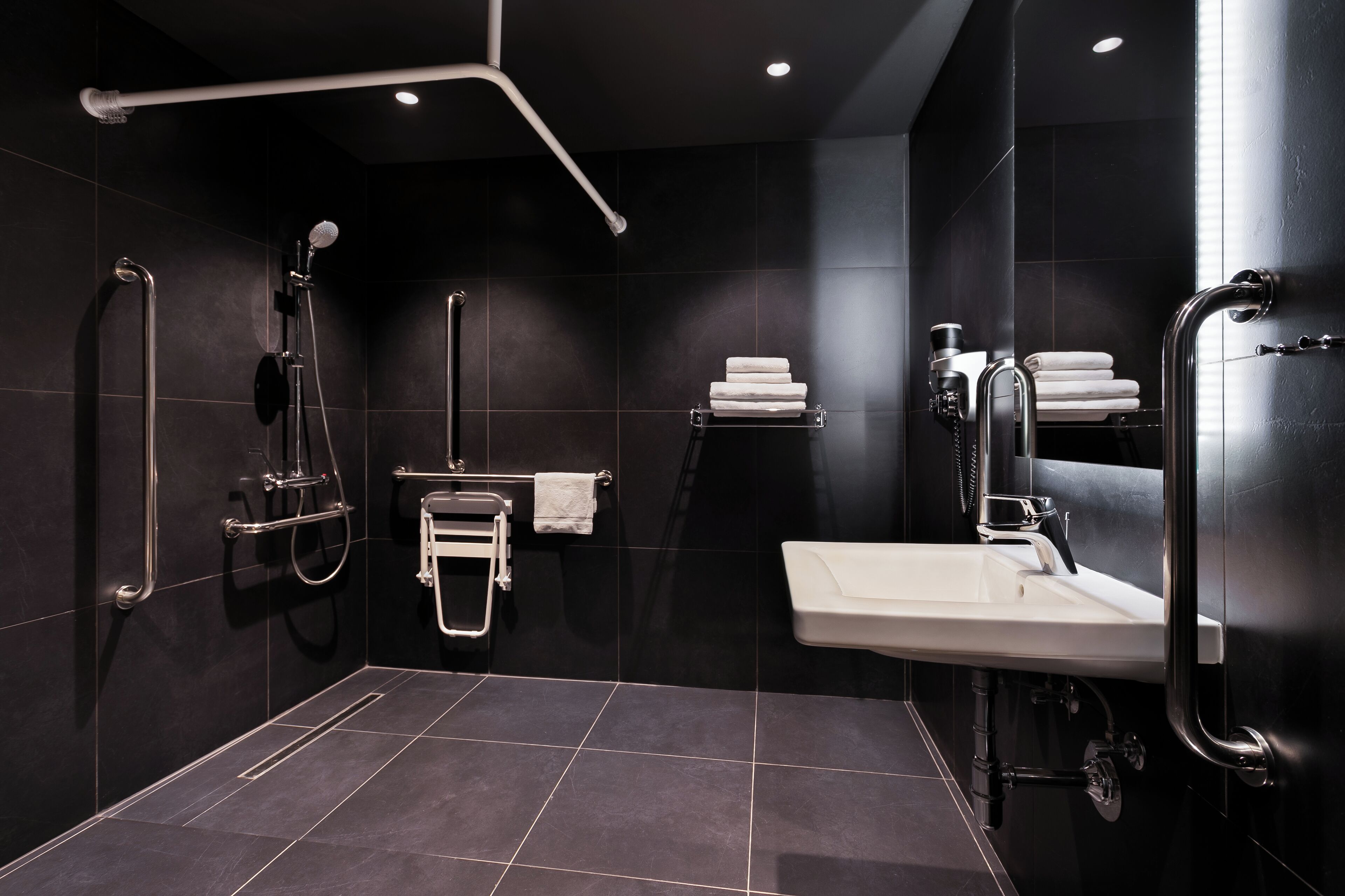 Ein modernes Badezimmer mit begehbarer Dusche, Haltegriffen, einem weißen Waschbecken und einem Handtuchhalter.
