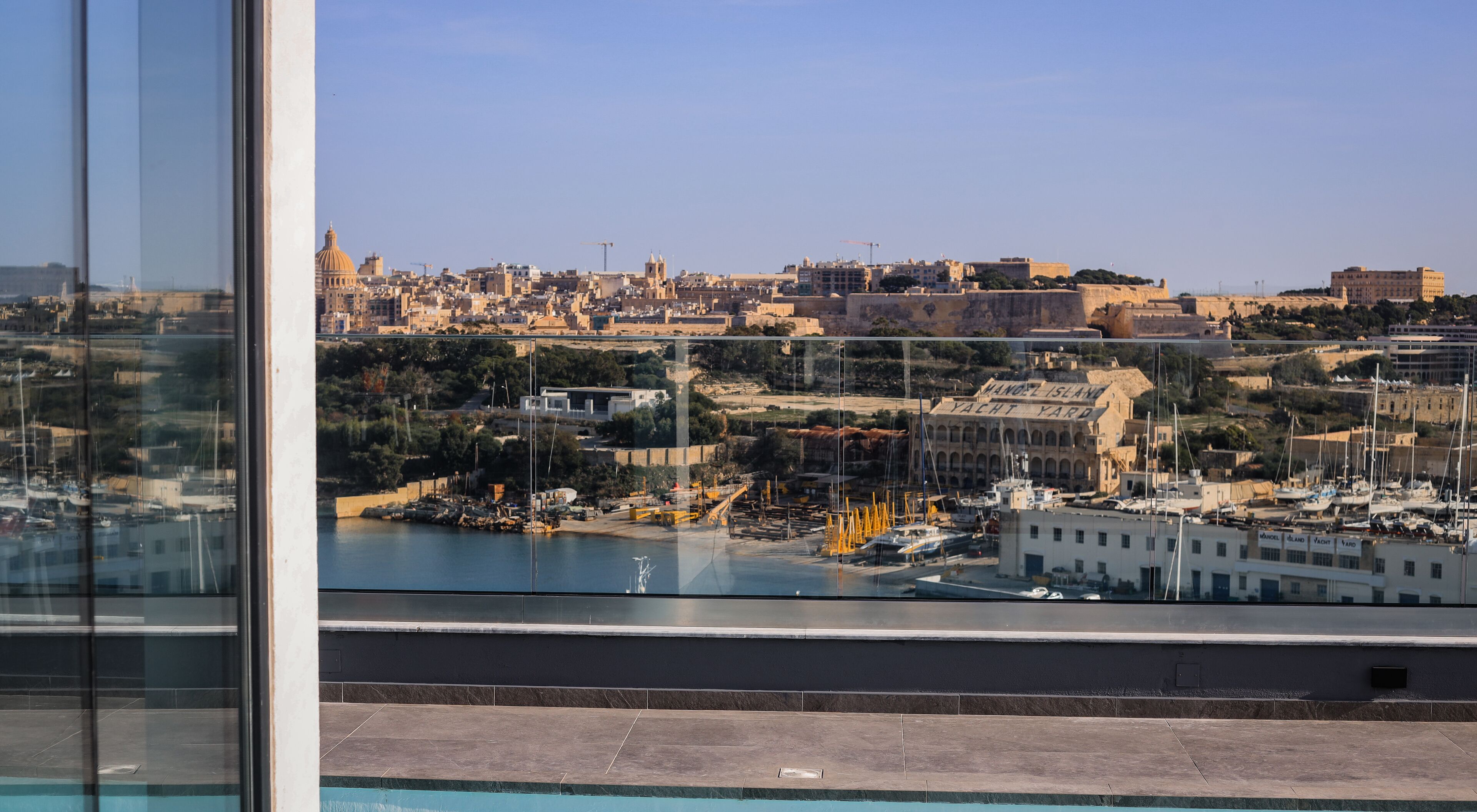 Blick von einem Balkon auf eine Marina mit Yachten, ein historisches Gebäude und eine Stadtlandschaft im Hintergrund.