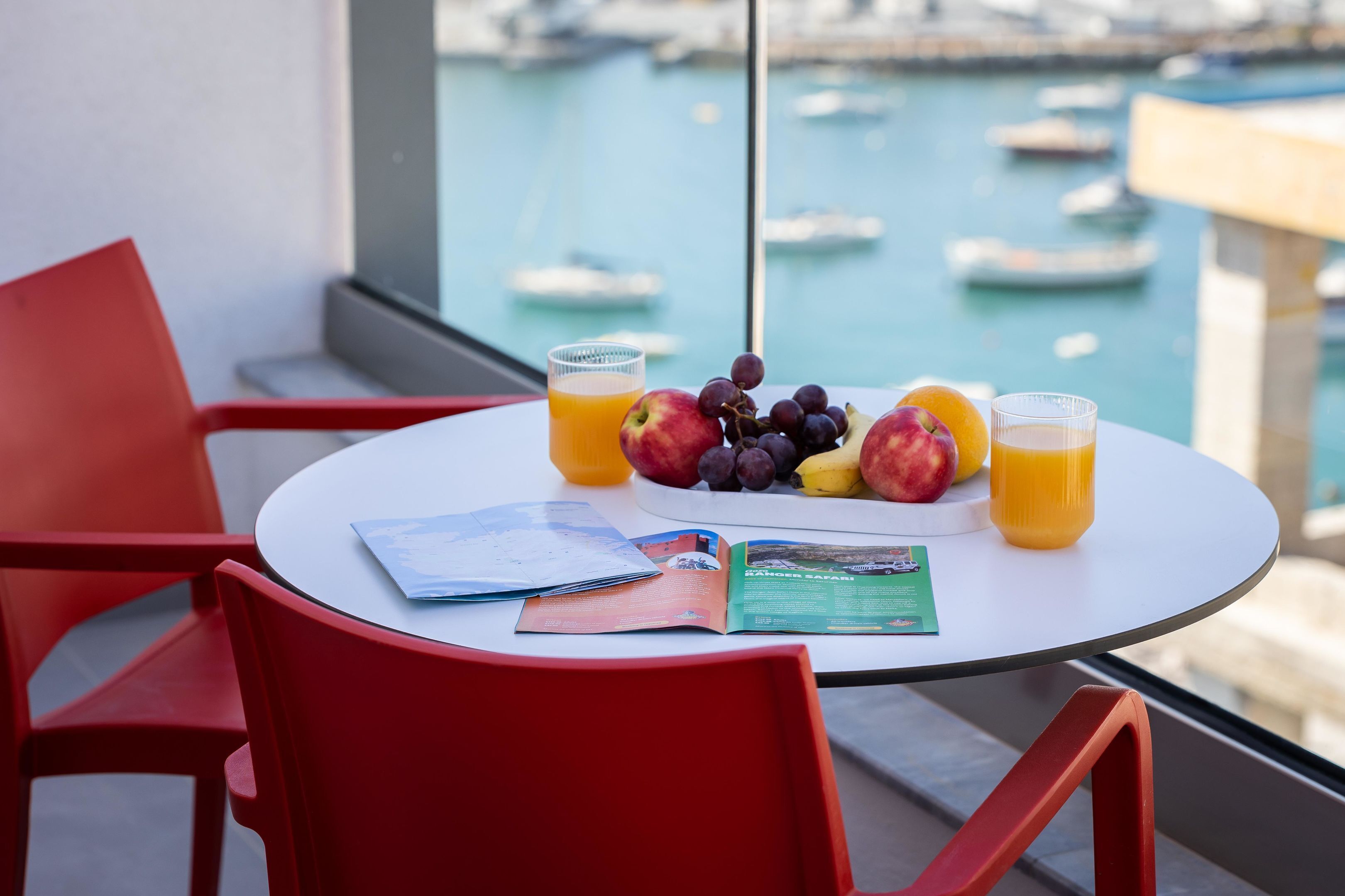 A table with a fruit plate, two glasses of orange juice, and open magazines on a balcony overlooking a marina.