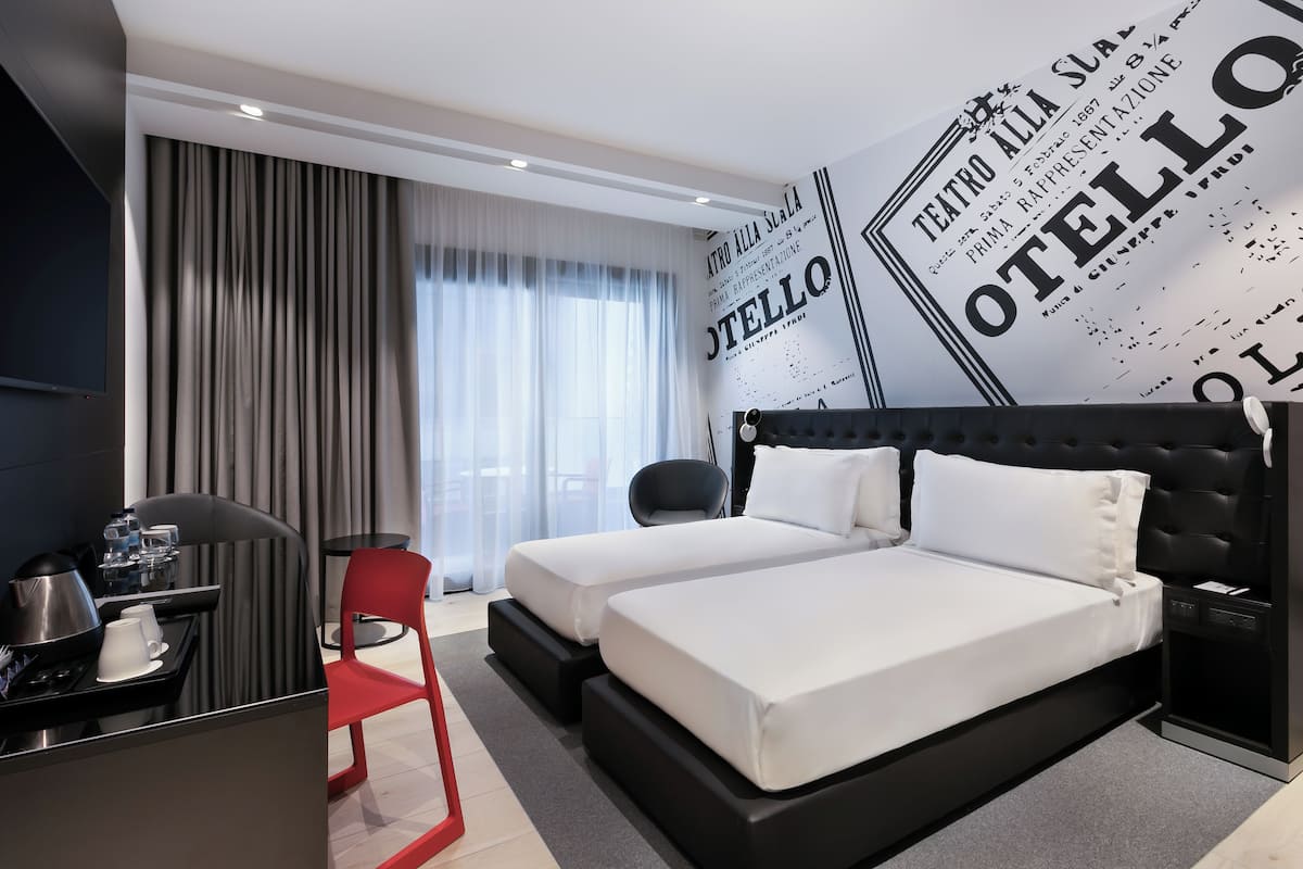 A hotel room with two beds, a desk with a coffee maker, a red chair, and a large window with curtains.
