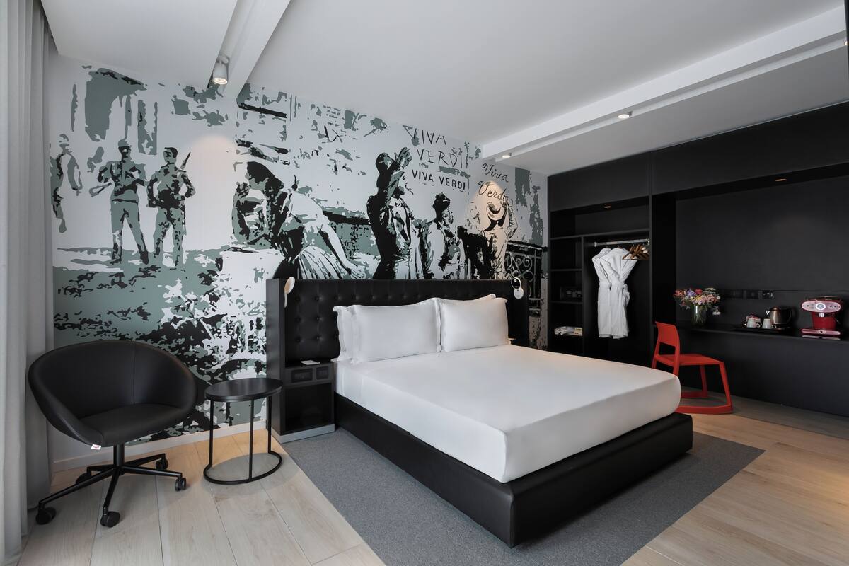 A modern bedroom with a large bed, a black office chair, a small side table, and a wardrobe.