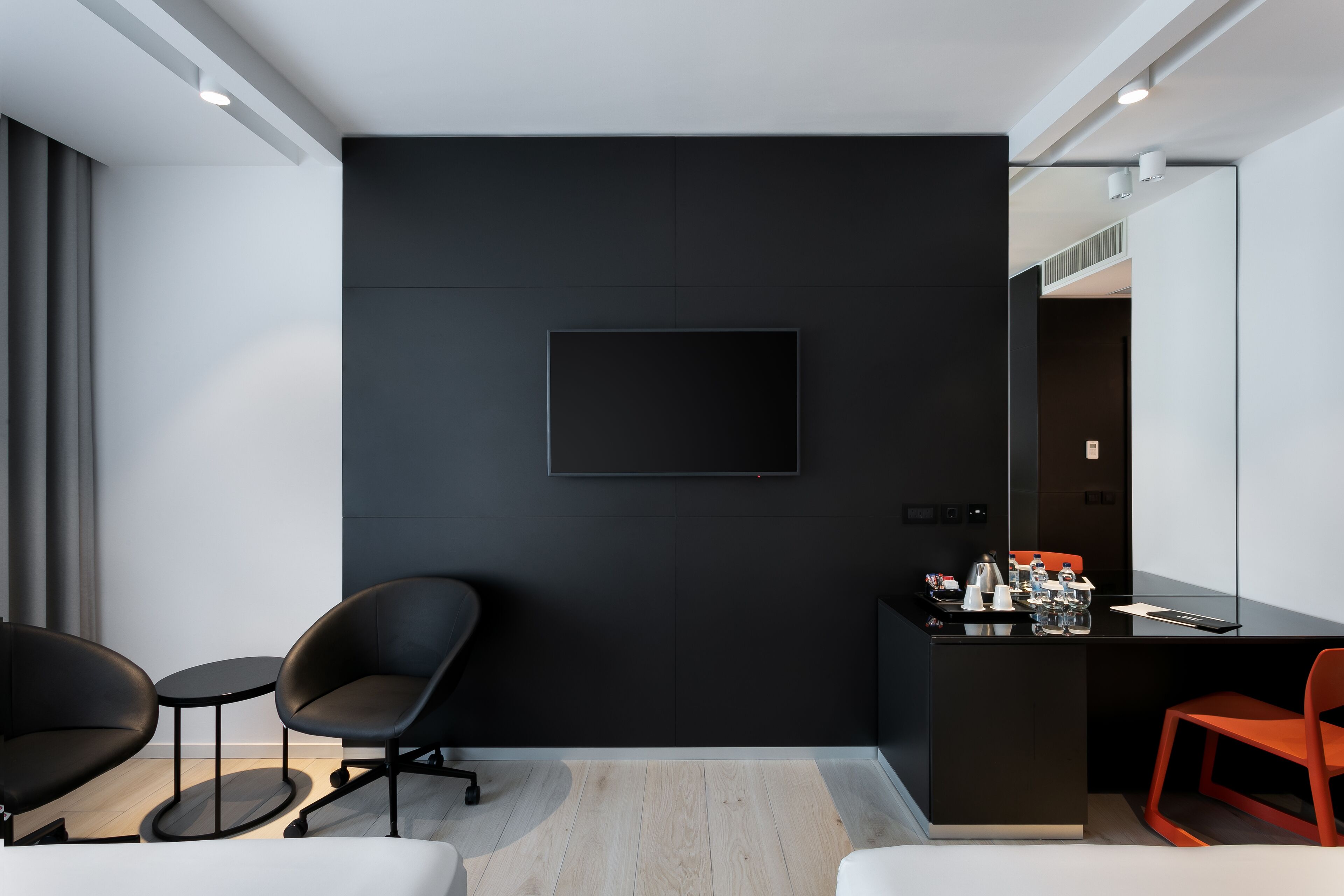 A modern hotel room with a black accent wall, a flat-screen TV, a small round table, two chairs, and a desk with a coffee machine.