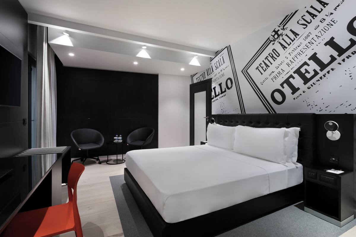 A modern hotel room with a large bed, two chairs, a desk, and a wall with a poster.