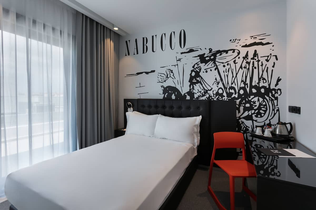 A hotel room with a large bed, a red chair, a desk with a kettle, and a wall mural with the word "NABUCCO".