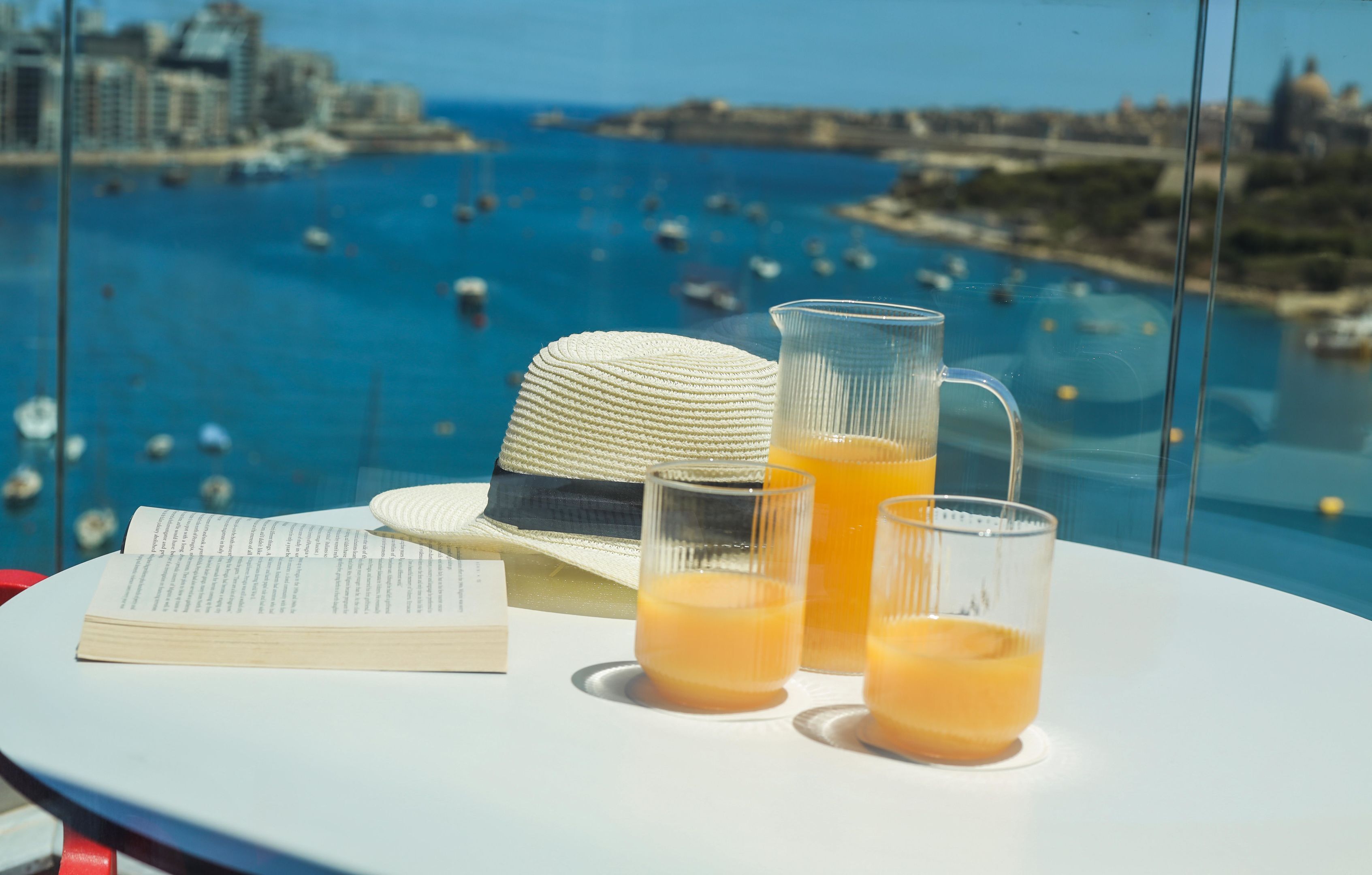 A table with a book, a straw hat, and two glasses of orange juice, overlooking a coastal cityscape with boats.