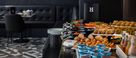 A buffet with various food items, including pastries and fruits, in a modern setting with a black leather couch and a geometric patterned floor.