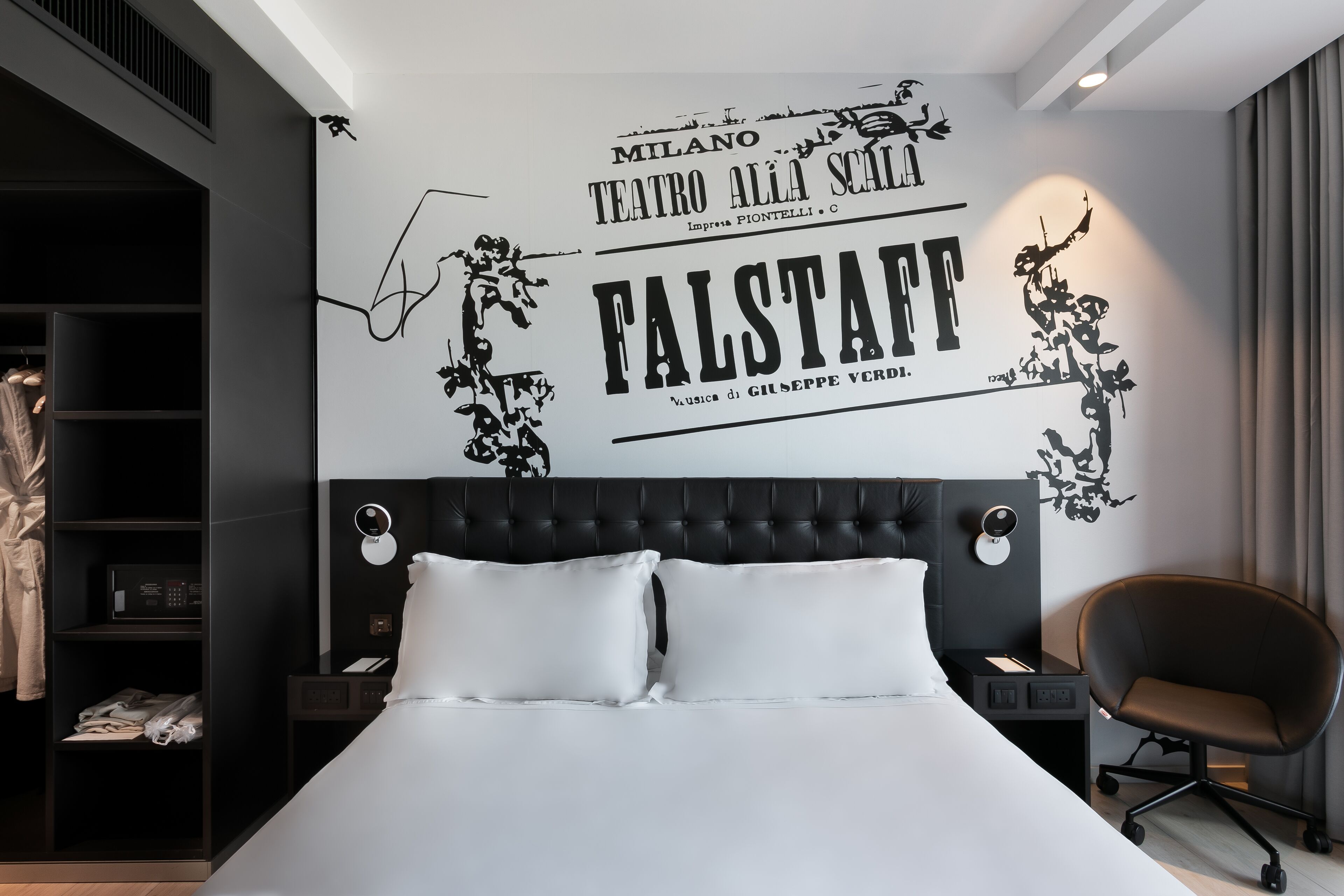 Et moderne hotelværelse med en stor seng, en sort sengegavl, en sort lænestol og en væg med et teaterplakat.