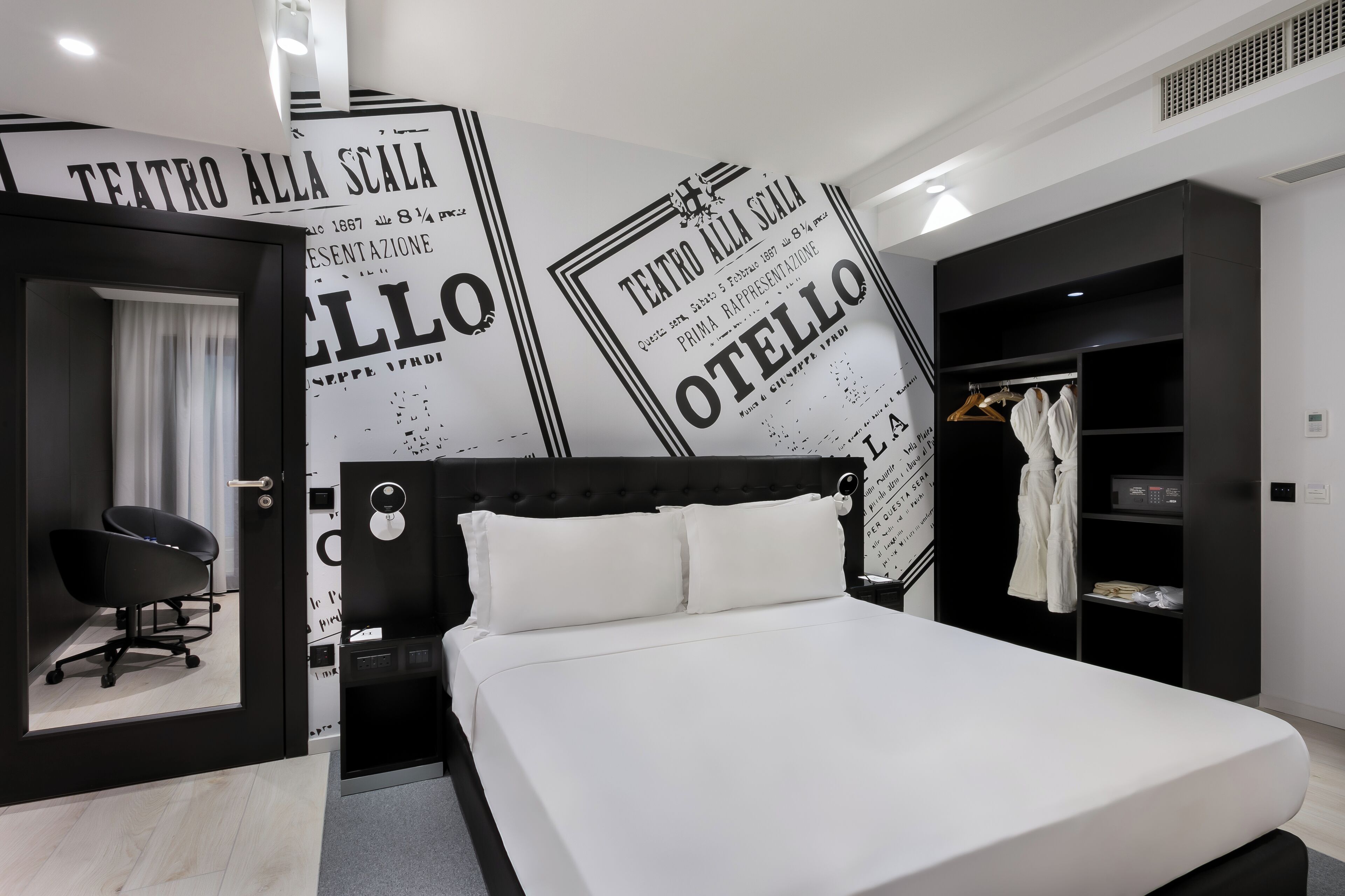 A modern hotel room with a large bed, a black office chair, a wardrobe, and a mirror.