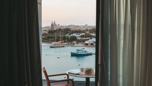 Vista desde un balcón con una mesa, una silla y un sombrero, que da al puerto con barcos y edificios.