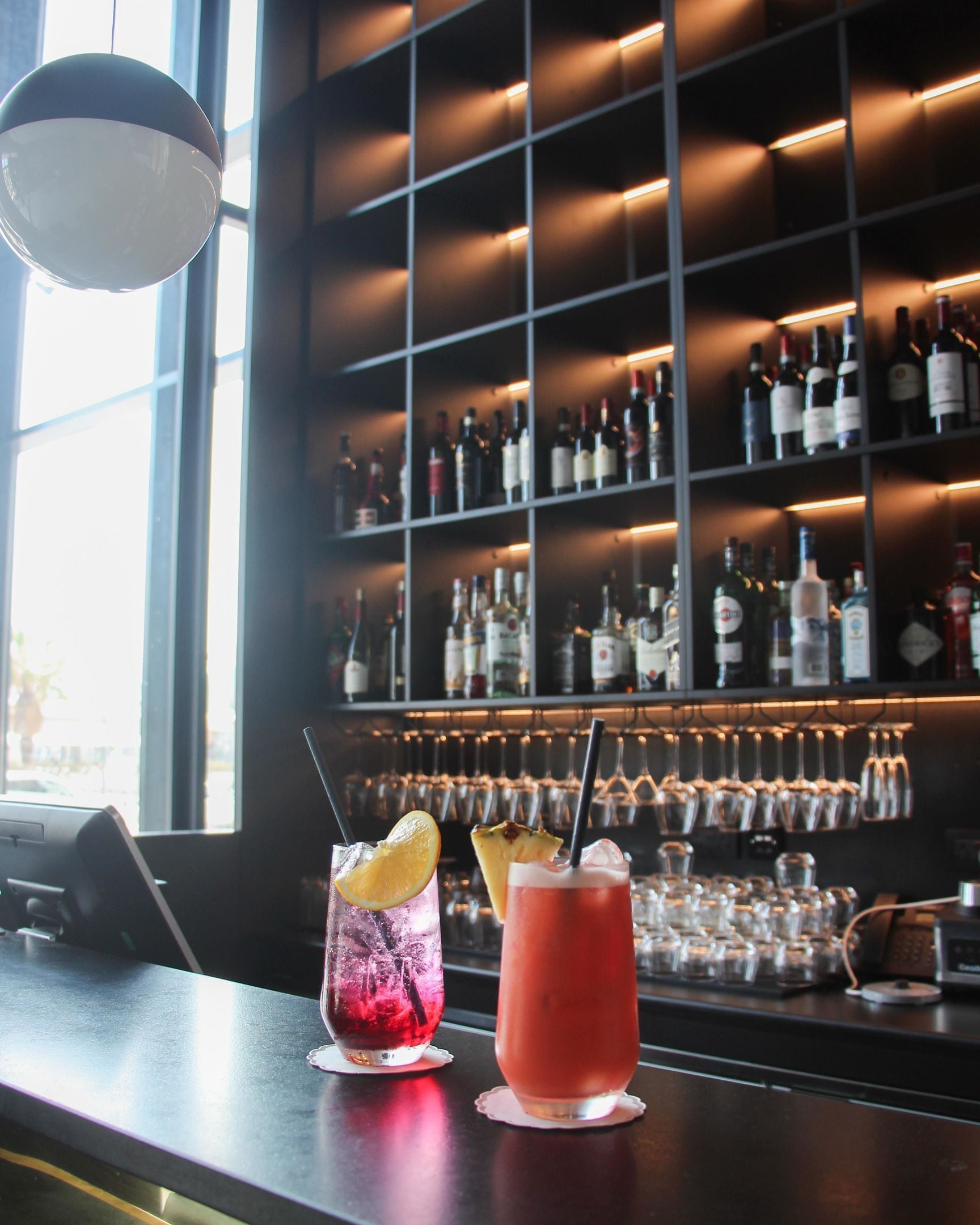 Una zona de bar con dos bebidas sobre la barra, una estantería para vinos al fondo y una lámpara colgante.