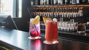 Una zona de bar con dos bebidas sobre la barra, una estantería para vinos al fondo y una lámpara colgante.