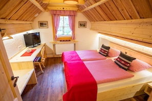 A cozy attic bedroom with a wooden ceiling, a desk with a computer, a bed with checkered bedding, and a window with red and white curtains.