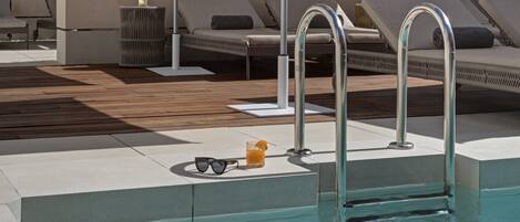 A poolside area with lounge chairs, a pool, and a small table with sunglasses and a drink.
