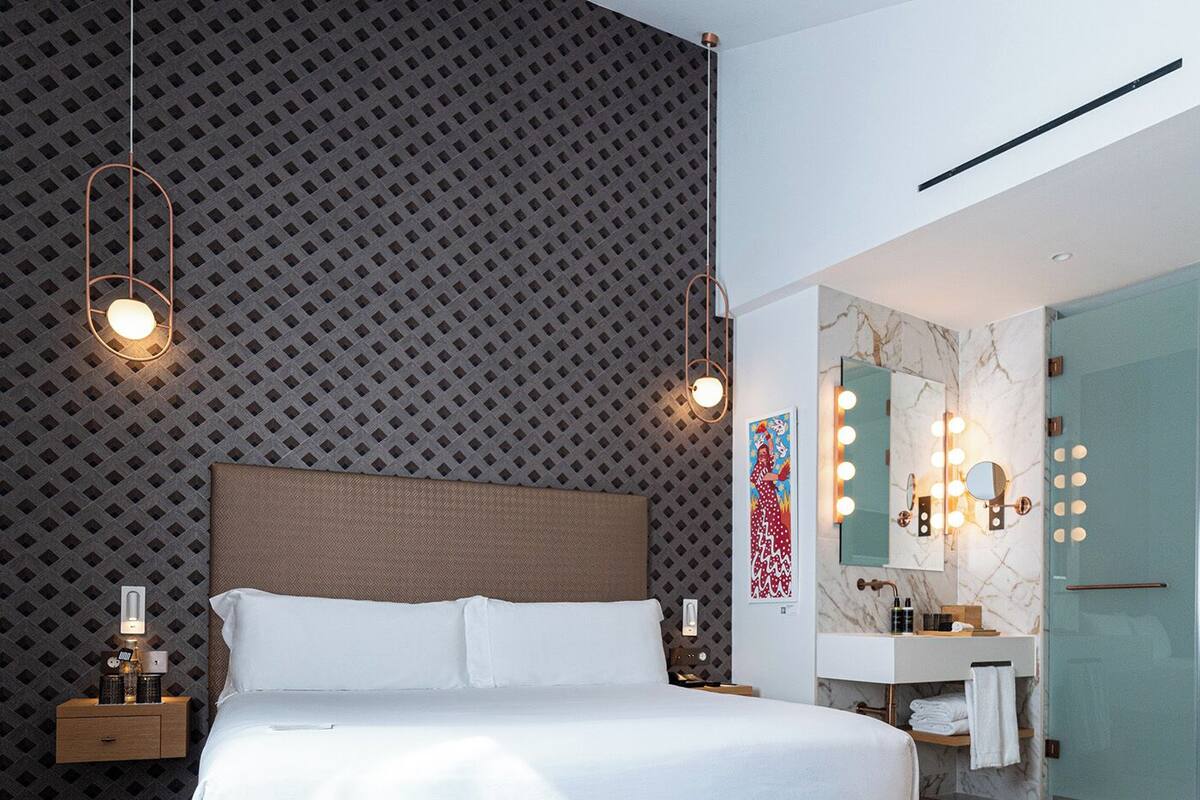 A modern hotel room with a large bed, two blue ottomans, a nightstand, a wall-mounted vanity with a mirror and lights, and a patterned wall.