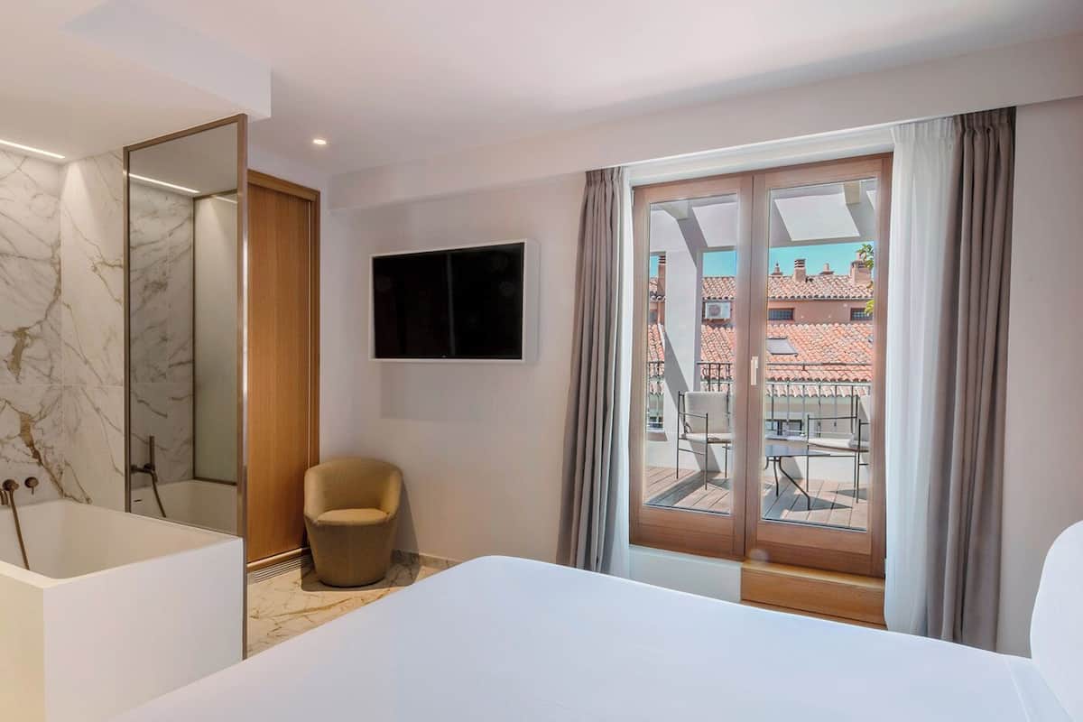 A modern hotel room with a large bed, a sitting area with a chair, a bathtub, a flat-screen TV, and a balcony with a view of buildings.