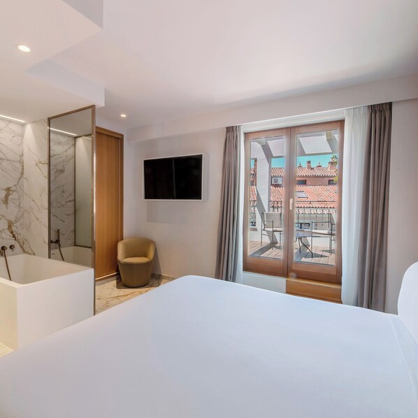 Una habitación de hotel moderna con una cama grande, un área de estar con una silla, una bañera, un televisor de pantalla plana y un balcón con vistas a edificios.