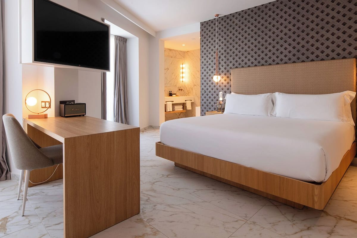 A modern hotel room with a large bed, a desk with a flat-screen TV, and a vanity with a mirror and lights.