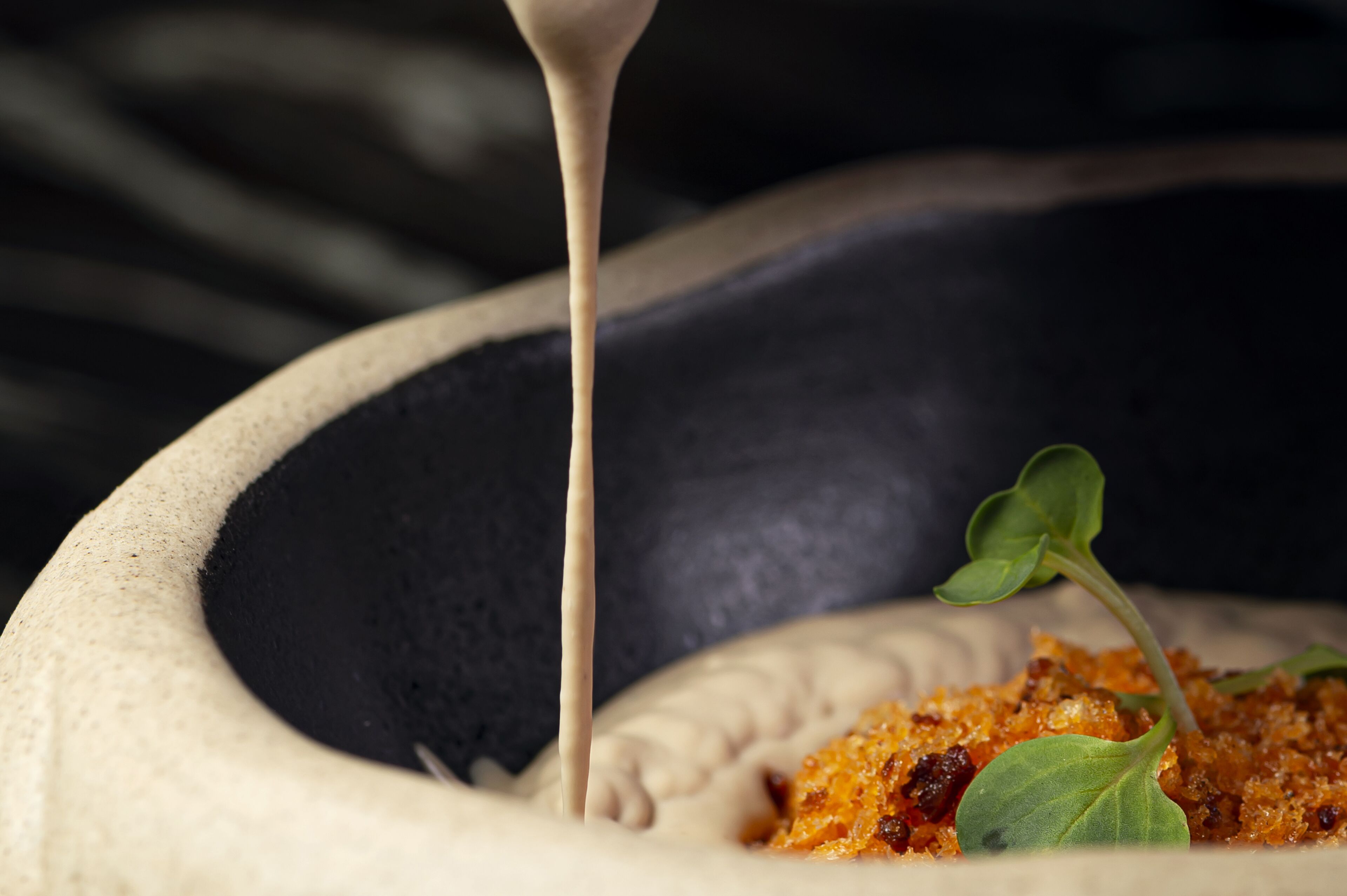 Primer plano de un plato con salsa cremosa que se vierte sobre un recipiente con un alimento desmenuzado, dorado y una pequeña guarnición de hoja verde.