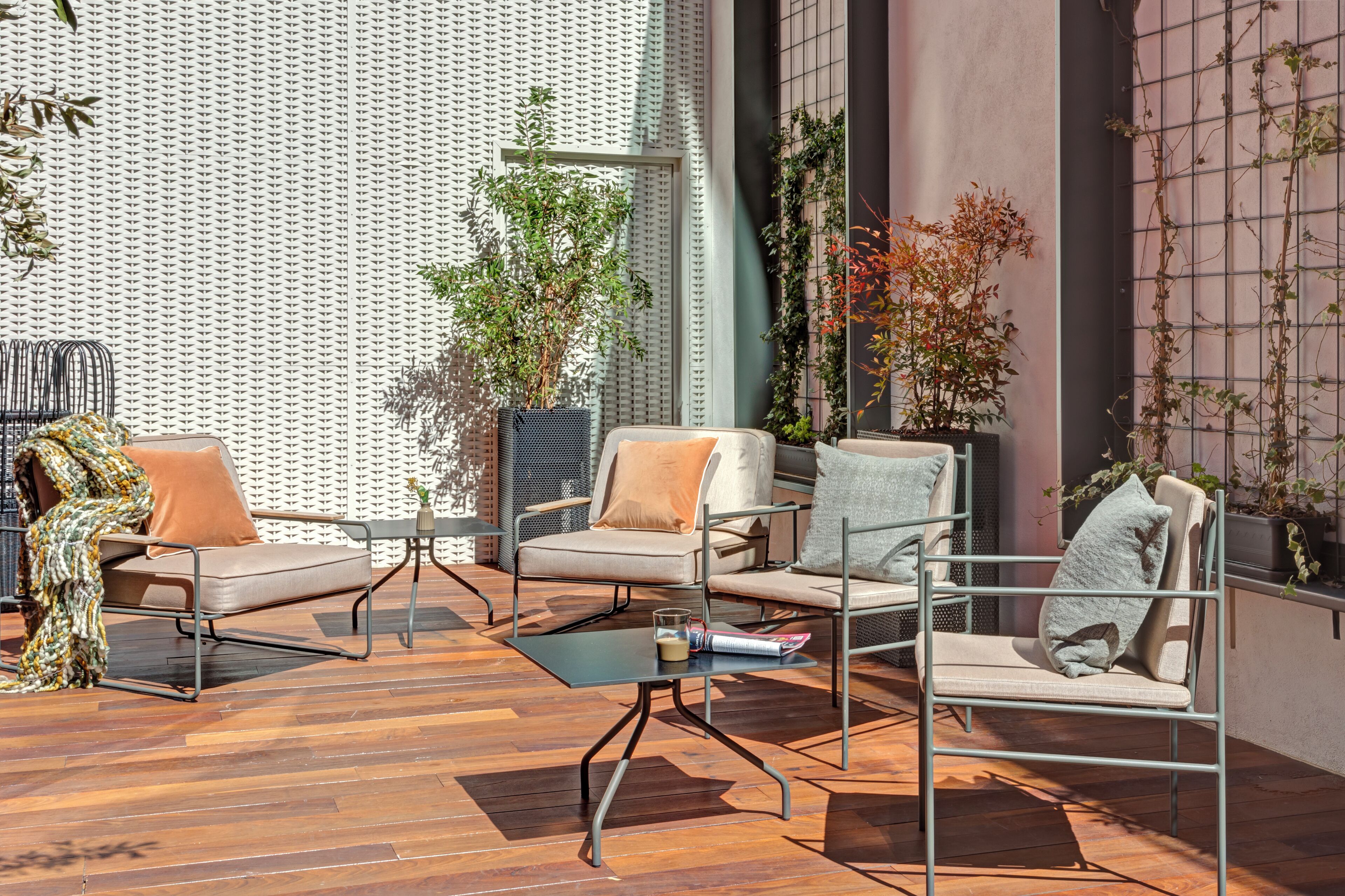Un espacio al aire libre con sillas de metal y una mesa de vidrio, rodeado de plantas en macetas y una pared con diseño.