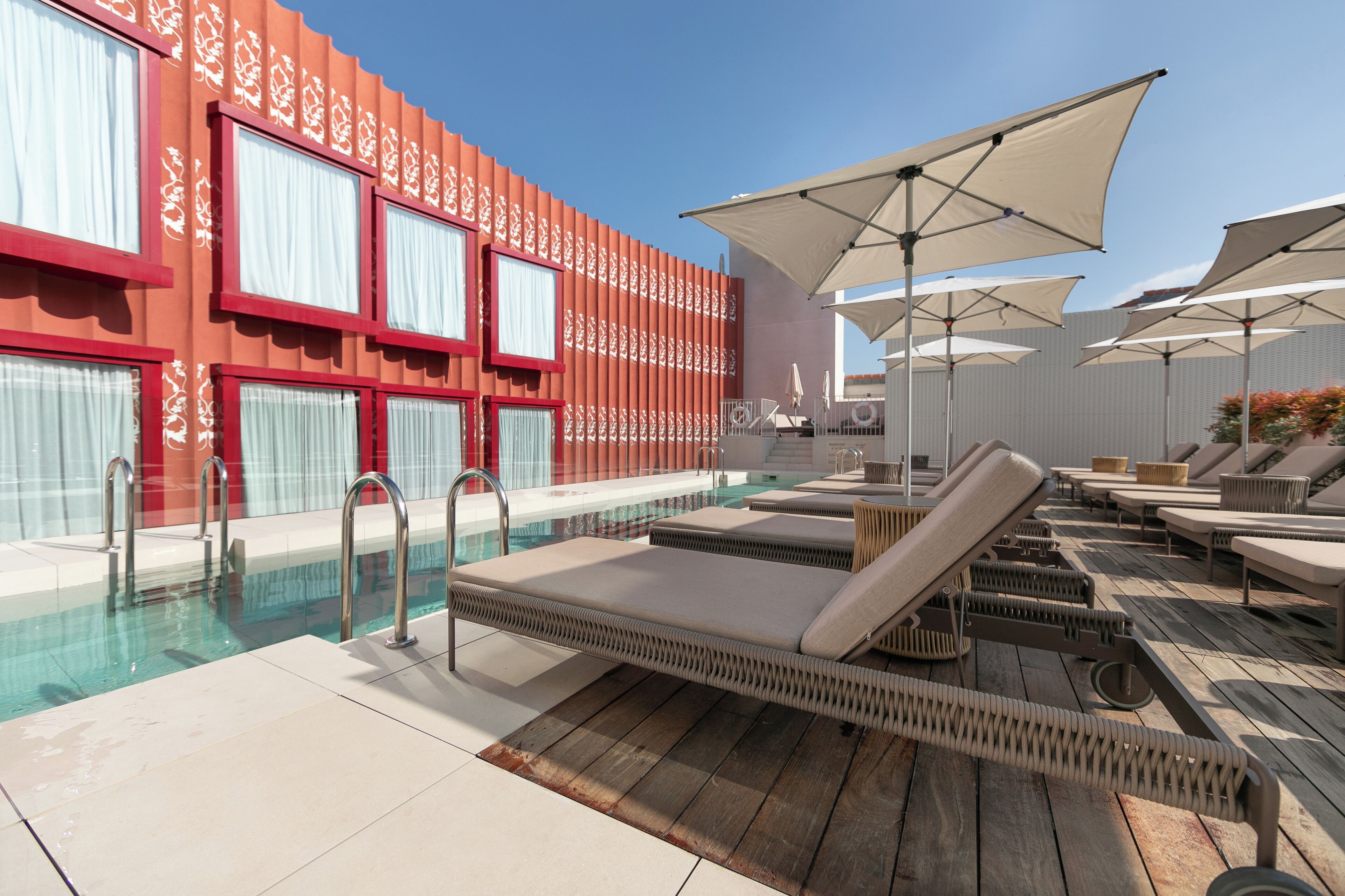 Zona de piscina con sillones reclinables, sombrillas y una pileta junto a un edificio con ventanas estampadas y detalles rojos.