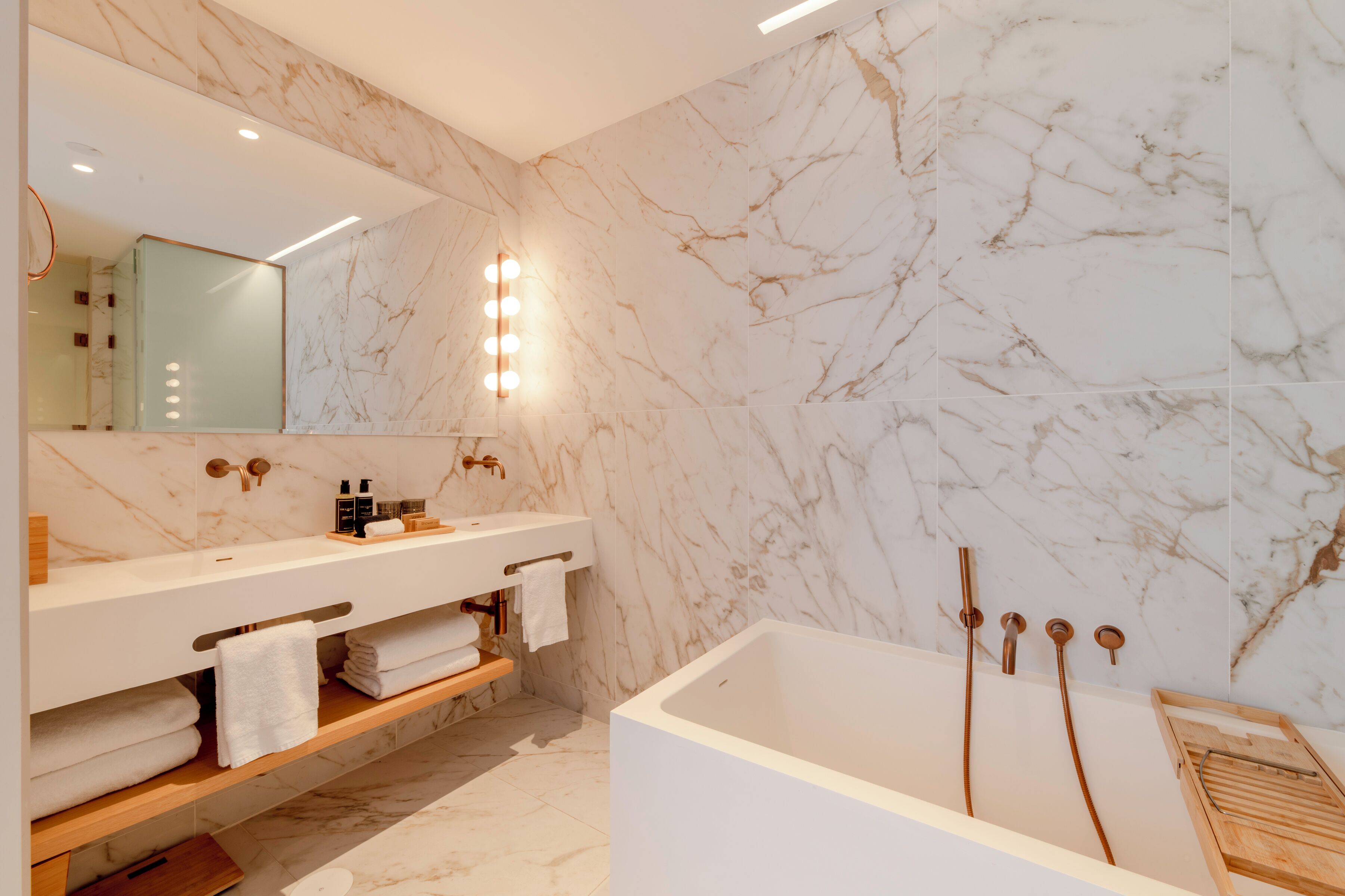 A modern bathroom with a marble wall, a large bathtub, and a vanity with a mirror and lights.