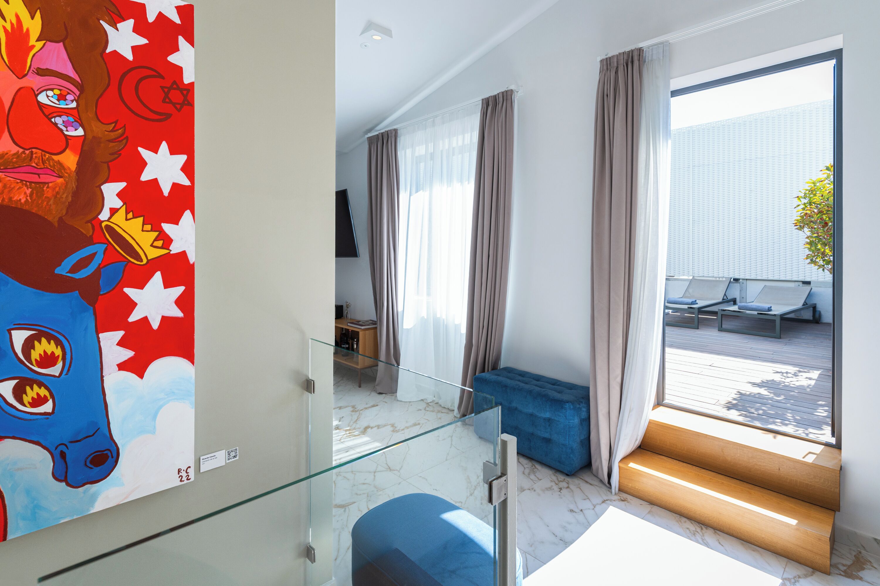 Habitación de hotel moderna con escalera de cristal, sofá azul y una ventana grande con cortinas.