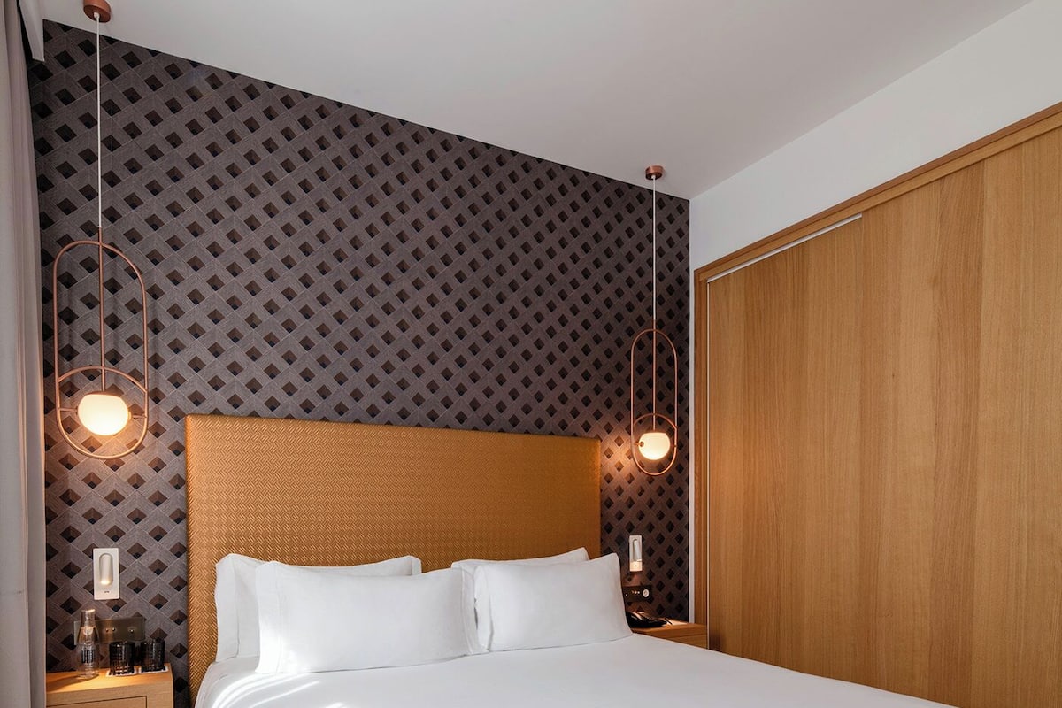 A hotel room with a large bed, wooden headboard, patterned wallpaper, and two bedside lamps.