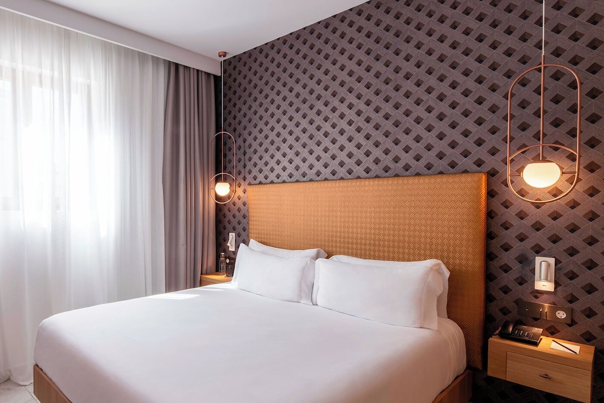 A modern hotel room with a large bed, a nightstand with a lamp, and a patterned wall.
