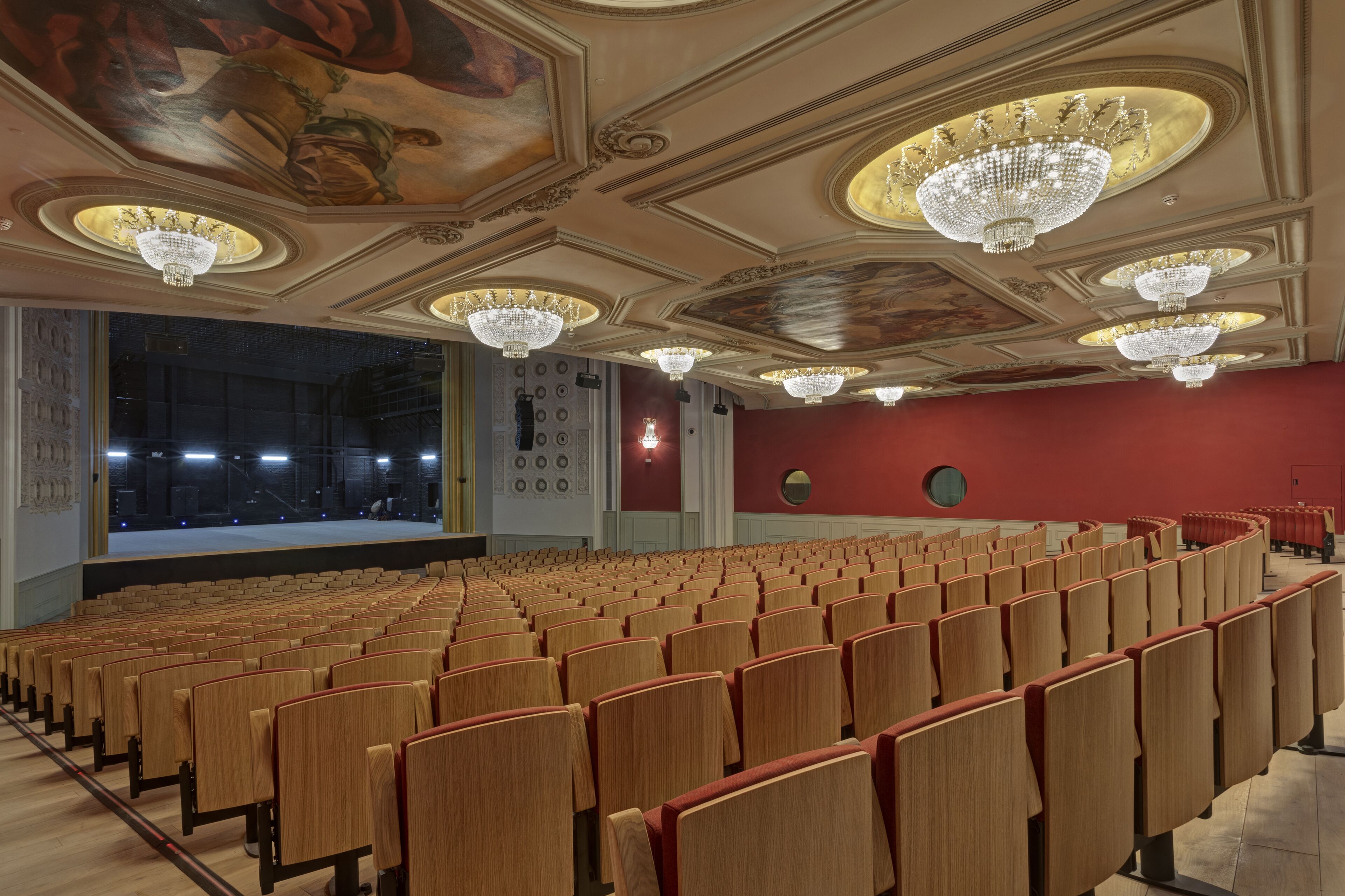 A theater with rows of wooden seats, ornate chandeliers, and a large mural on the ceiling.