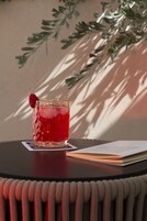 Een glas rode drank met een framboos op een onderzetter, een boek en een plant op de achtergrond.