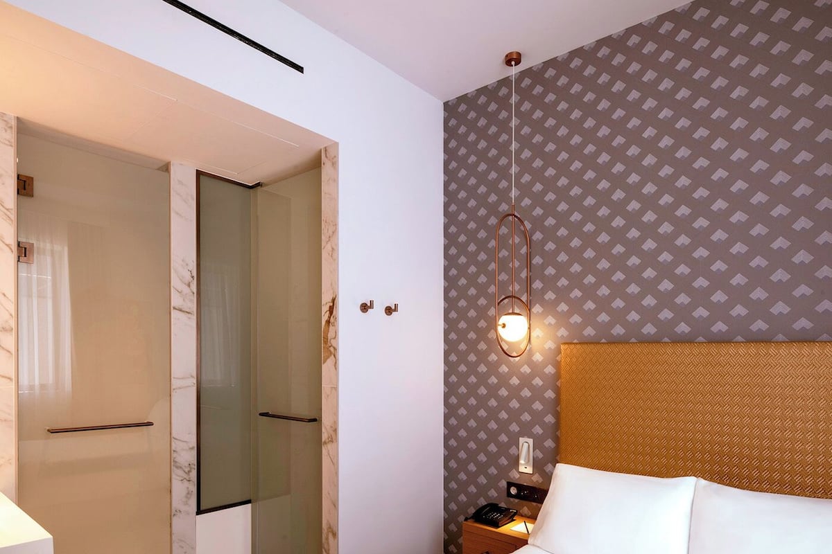 A hotel room with a bed, a nightstand with a lamp, a mirror, and a door leading to a bathroom.