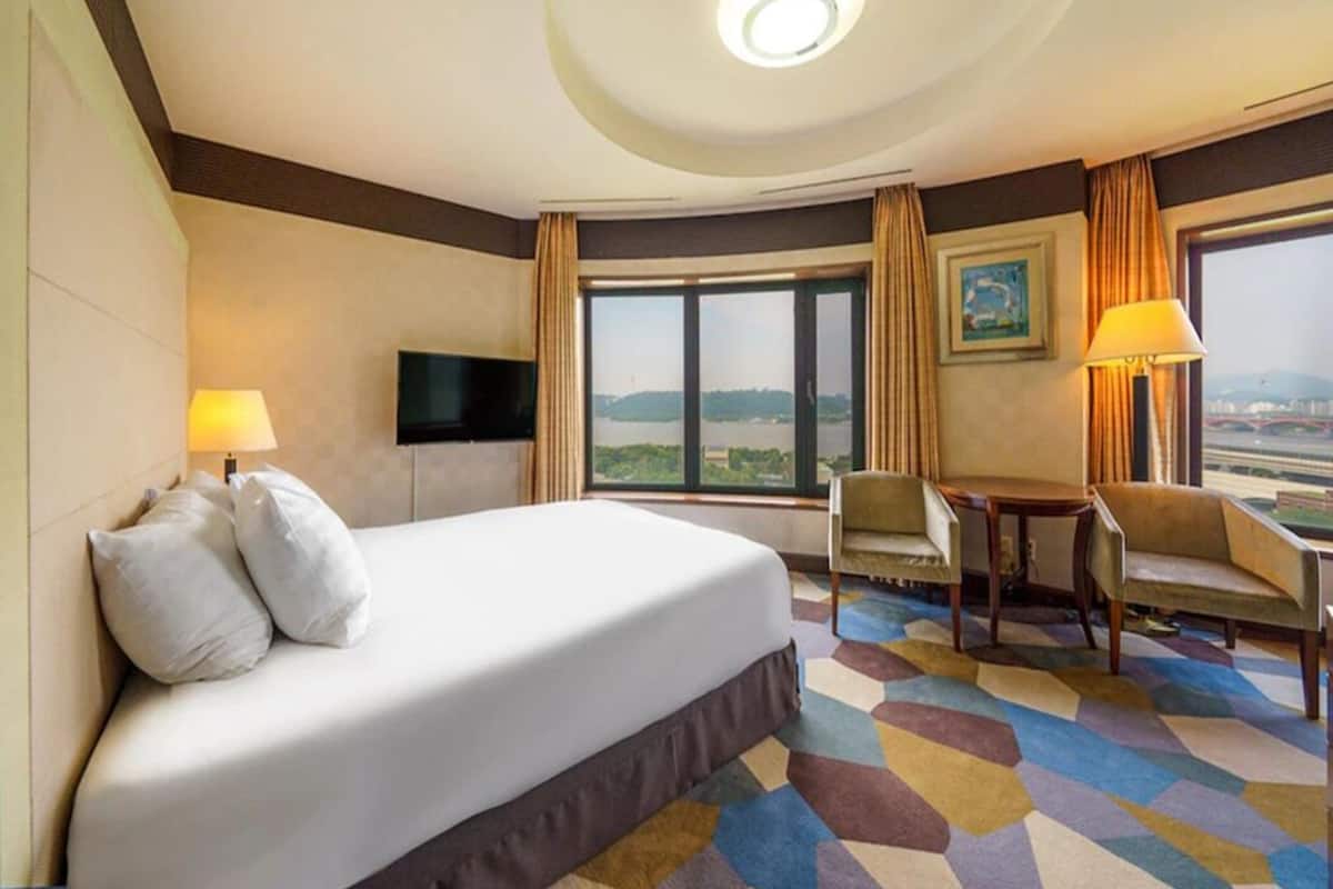 A hotel room with a large bed, a flat-screen TV, a desk with a chair, and a view of the cityscape through large windows.