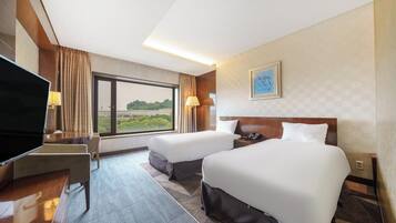 A hotel room with two beds, a desk with a TV, and a large window offering a view of greenery.