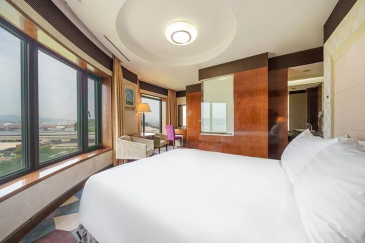 Ein Hotelzimmer mit einem großen Bett, einem Schreibtisch, einem Sessel und Blick auf die Skyline durch große Fenster.